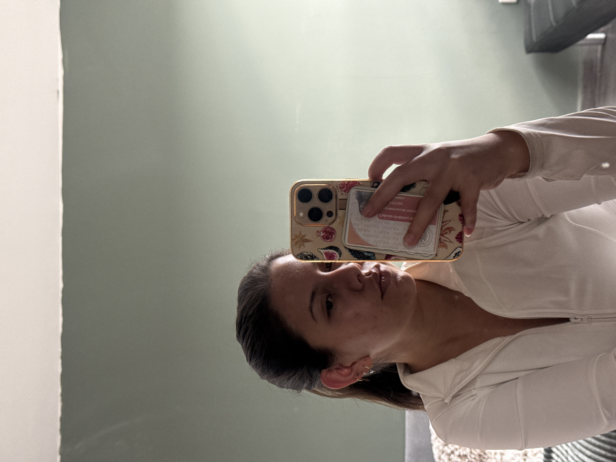 An indoor scene in San Bernardino Tlaxcalancingo, Mexico, depicts a female individual taking a mirror selfie. The subject is a young adult female with dark hair pulled back, wearing a white long-sleeved top with a partially unzipped collar. Her left ear is adorned with multiple small earrings. She holds a gold-colored smartphone in her right hand, visible in the reflection, positioned to capture her upper body and face. The phone case features a decorative pattern of pomegranates or figs and a star design, with a cardholder attached containing a card with red text.
The background consists of a muted green wall with a white ceiling line visible at the top. To the left, a light-colored upholstered object, possibly a sofa or bed, is partially visible, along with a patterned blanket. To the right, a dark gray or black rectangular furniture item with metallic legs is present. The lighting appears to be natural, entering from the left, illuminating the subject and creating a subtle gradient on the green wall.