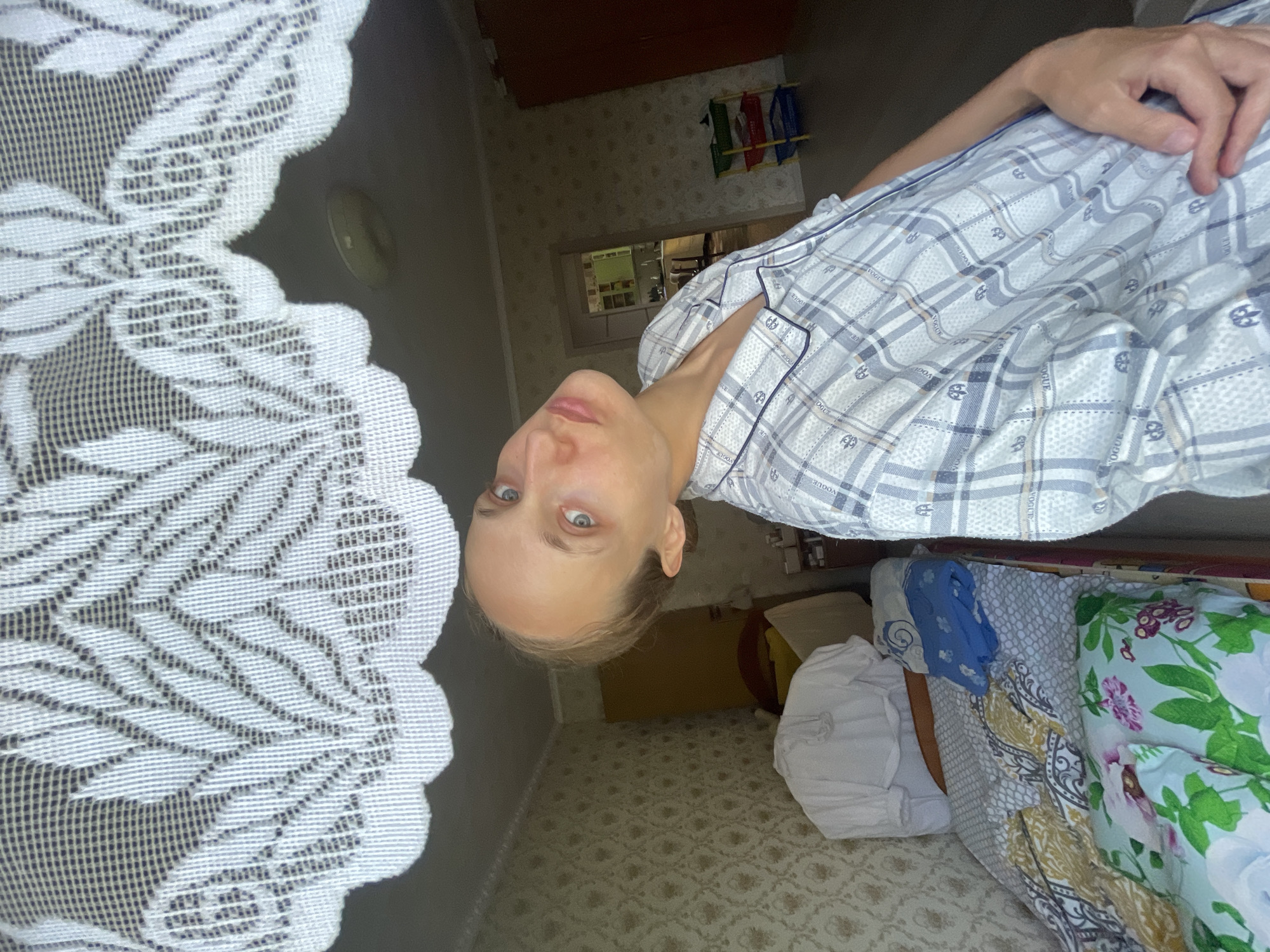 The image shows a woman in a room. The room appears to be a bedroom or a living space, with a bed, a wardrobe, and wallpaper on the walls. There is a white lace curtain hanging in the foreground, partially obscuring the view of the room. The woman is wearing pajamas and is looking towards the camera with her head tilted. The lighting suggests it is daytime, and the overall atmosphere is casual and domestic. The image has been taken from a low angle, looking upwards towards the woman. There are no specific details in the image that would allow for identifying the exact location within the city of Sharkan, Russia.
