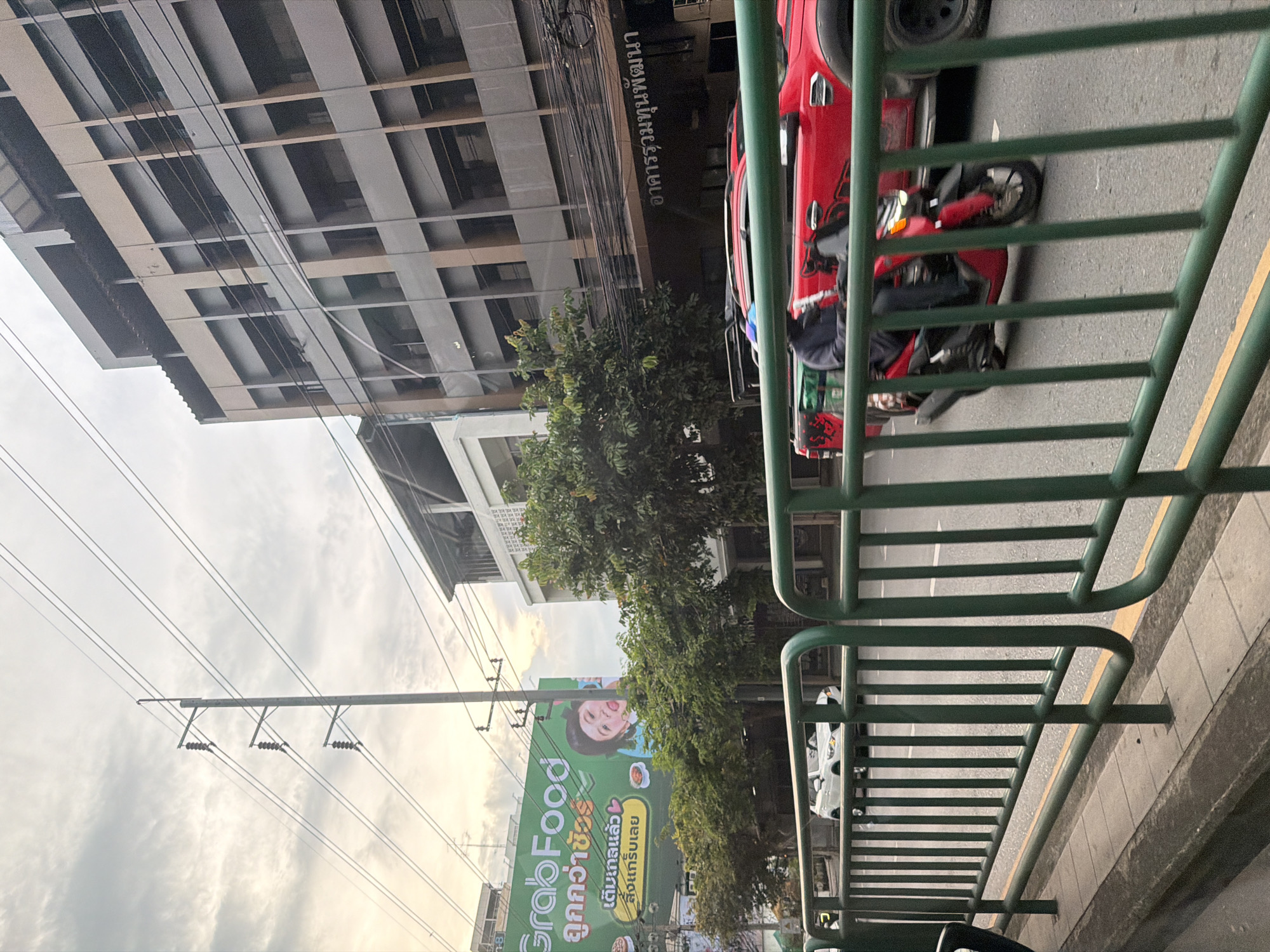 The image depicts a street scene in Bangkok, Thailand, likely during daylight hours with overcast skies. In the foreground, a green metal railing lines the sidewalk. Beyond the railing, traffic moves along a paved road. A red scooter with a rider wearing a blue helmet and dark clothing is prominently visible in motion. To the right, the rear portion of a red pickup truck is also seen. Further back, a white SUV is visible on the road. On the left side of the image, a large green billboard advertises "GrabFood" with Thai text and an image of a child. Behind the billboard, trees are visible, and beyond them, multistorey buildings rise. One building on the right, appearing to be an office building, has a dark facade with rectangular windows. Electrical poles and numerous overhead wires traverse the sky, typical of urban infrastructure in Bangkok. The overall impression is a busy urban environment.
