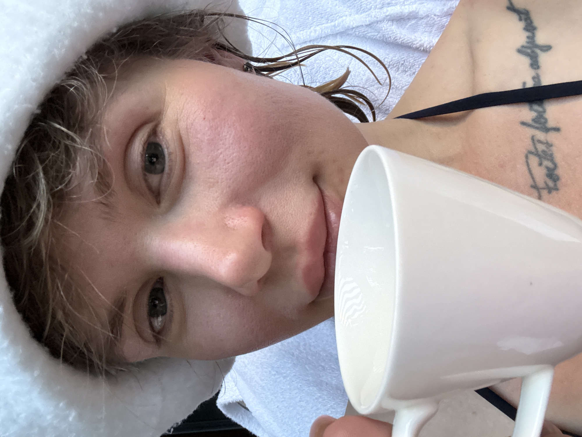 A close-up selfie shows a woman wearing a white towel headdress and a black bikini top. She is holding a white mug with both hands, bringing it up to her mouth. Her hair is wet and falls around her face and shoulders. A tattoo is visible on her upper chest. The background consists of white towels. The image was taken indoors, likely in a spa or bathroom setting, but the specific location within Urlu-Aspak, Russia, cannot be determined from the visual information provided.