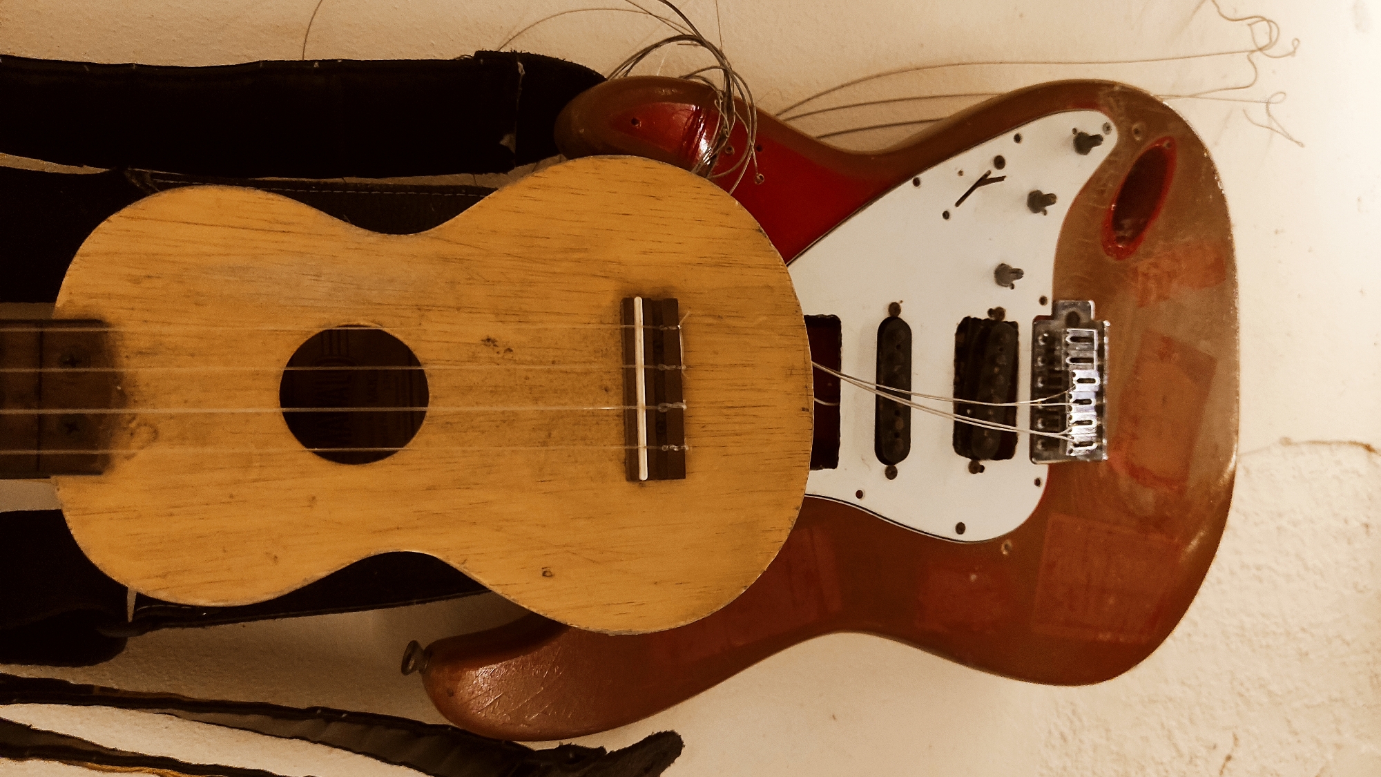 The image displays a close-up view of two stringed instruments, likely a ukulele and an electric guitar, hanging on a textured wall. The ukulele, made of light-colored wood, is positioned in front of the guitar. It features a circular soundhole and a wooden bridge with four strings. The electric guitar, with a red-brown body and a white pickguard, is visible beneath the ukulele. Its strings are detached from the ukulele and loosely hang, some coiled and others trailing down. The background wall is off-white and shows a rough texture. The lighting is warm, casting soft shadows. The scene appears to be an indoor setting, potentially a storage area or a music room. There are no people or visible actions taking place.
