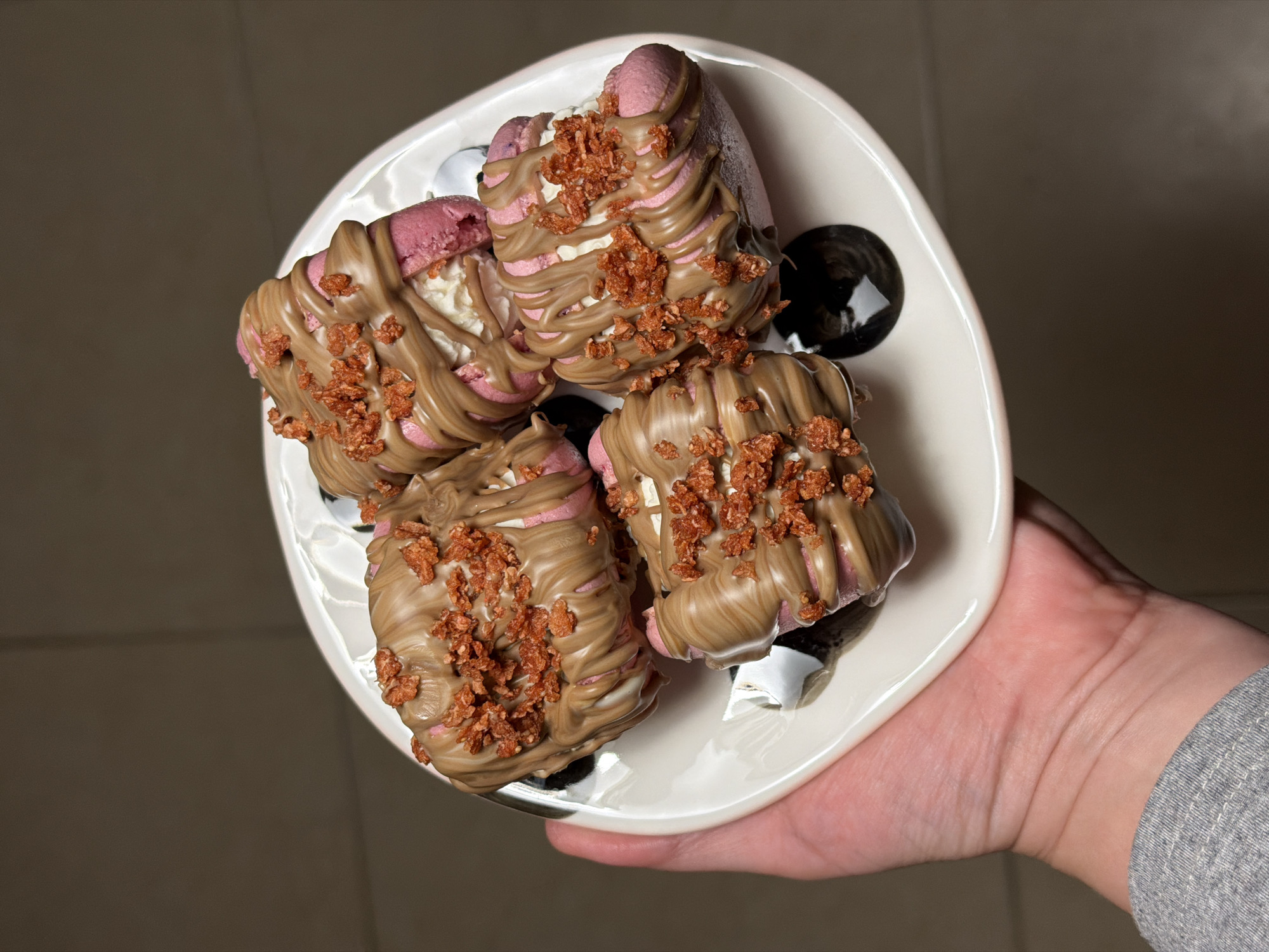 A top-down, slightly angled shot shows a hand holding a white, irregularly shaped ceramic plate laden with four dessert items. The desserts appear to be large, pink-colored macarons or meringues, filled with a white creamy substance. Each dessert is generously drizzled with a light brown, glossy sauce, resembling milk chocolate or caramel, and topped with a scattering of reddish-brown, crunchy bits, possibly granola, nuts, or cookie crumbles. The plate features subtle black decorative elements in its recesses. The hand holding the plate is visible in the lower right corner of the frame, wearing a grey long-sleeved top, possibly a sweater. The background is a plain, dark taupe or warm brown color, suggesting an indoor setting. The lighting is artificial, illuminating the desserts and plate clearly. No text is visible in the image.