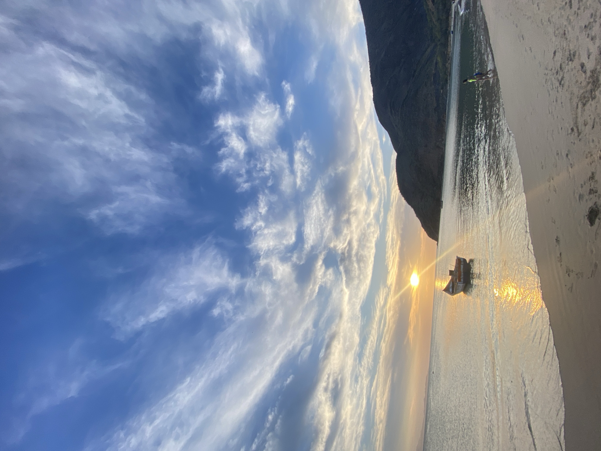 This is a landscape photograph of Juan Griego, Venezuela. The scene depicts a beach with a small boat resting in the water near the shoreline. The sun is setting on the horizon, casting a golden reflection on the sea. A person is standing in the water. Mountains form the backdrop of the scene. The sky is filled with clouds, suggesting either sunrise or sunset.