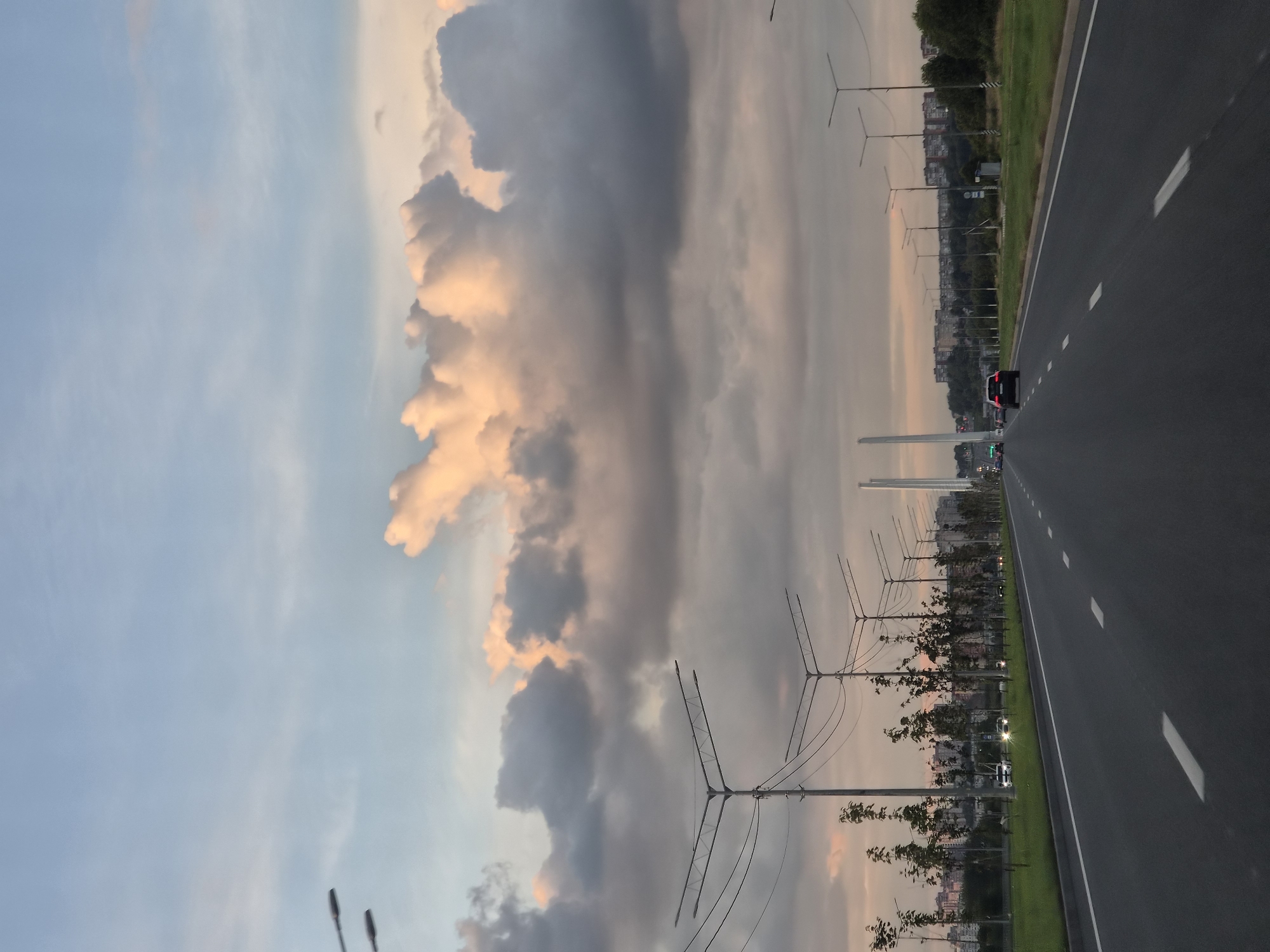 The image shows a road with streetlights and trees lining the sides. There are several cars on the road, and buildings can be seen in the background. The sky is filled with clouds, and the sun is setting. The scene appears to be a highway in Cherepovets, Russia.