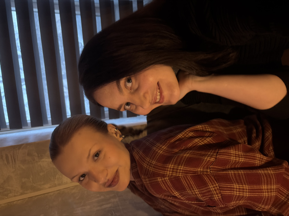 Two young adult women are depicted in an indoor setting in Chaikovsky, Russia. The woman on the left has light-colored hair styled back, fair skin, and wears a red and yellow plaid shirt. She is positioned facing slightly right of the camera, with her head tilted, displaying a subtle smile and looking directly forward. Her visible left ear has two small hoop earrings. The woman on the right has shoulder-length dark hair, fair skin, and is wearing a dark, possibly black, top. She is seated slightly behind and to the right of the first woman, smiling broadly and looking towards the camera, with her right hand resting under her chin or jawline. The background features a light-colored, textured wall on the left and a window with horizontal blinds on the right. The scene is illuminated by warm, soft lighting, suggesting an evening or indoor ambiance.
