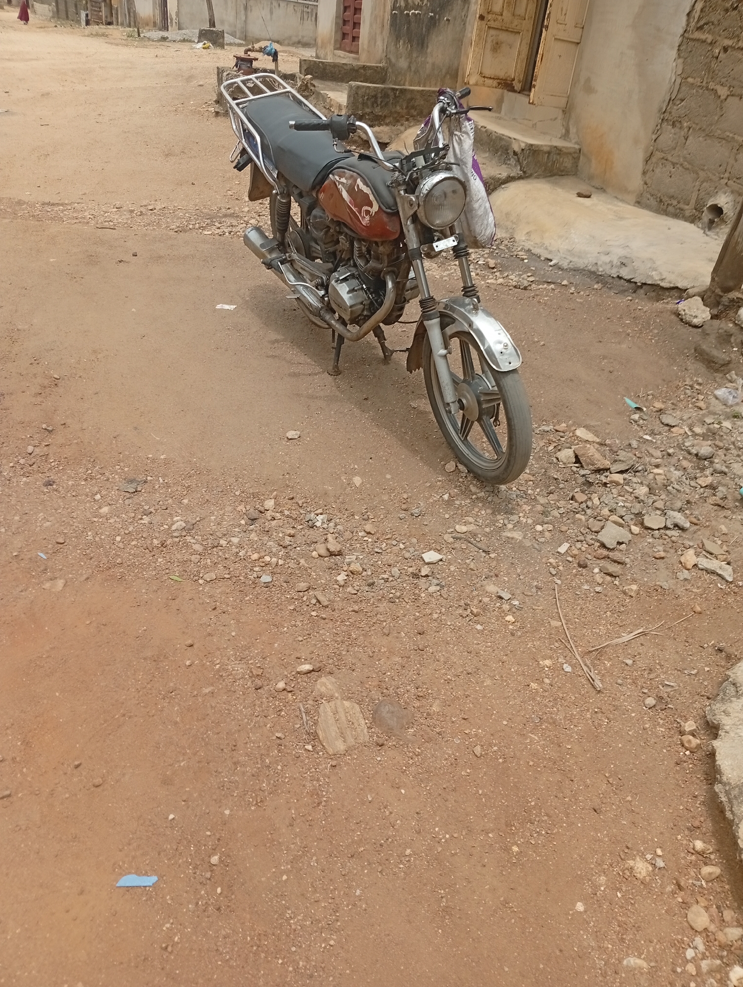 The image shows a motorcycle parked on a dirt road in Adankolo, Nigeria. The motorcycle is brown and silver with a black seat. It is facing slightly to the right of the frame. The road is unpaved, composed of dirt and gravel. In the background, there are buildings, one with an open door and steps. A white bag hangs on the left handle bar. The overall lighting suggests a sunny day.