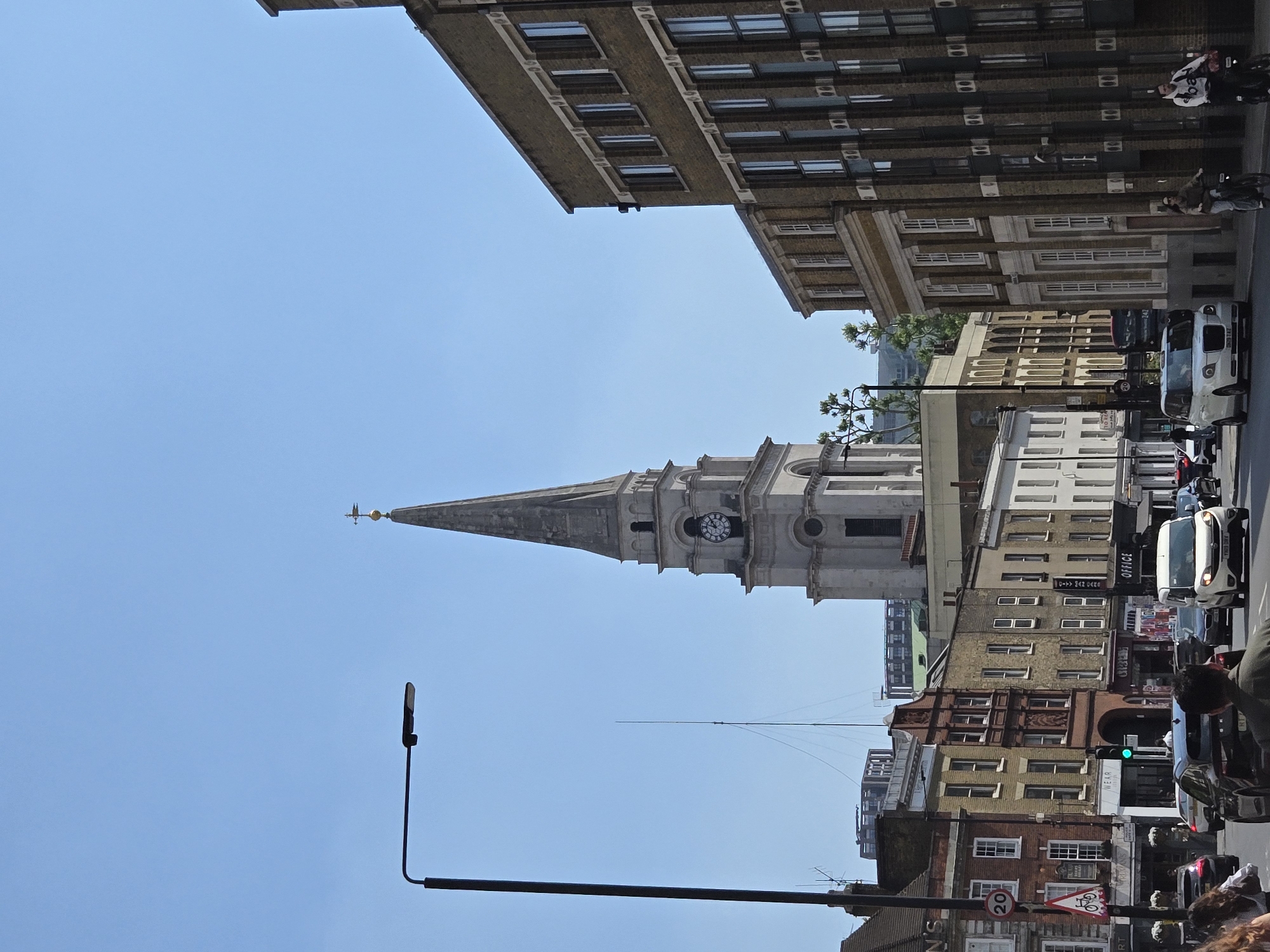 The image shows a street scene in London, United Kingdom. A tall church spire dominates the view, rising above buildings. Buildings of varying heights and architectural styles line the street. Vehicles, including cars and vans, occupy the street. Pedestrians are walking along the pavement. A lamppost stands in the foreground. The sky is a clear blue. A road sign indicates a 20 mph speed limit.