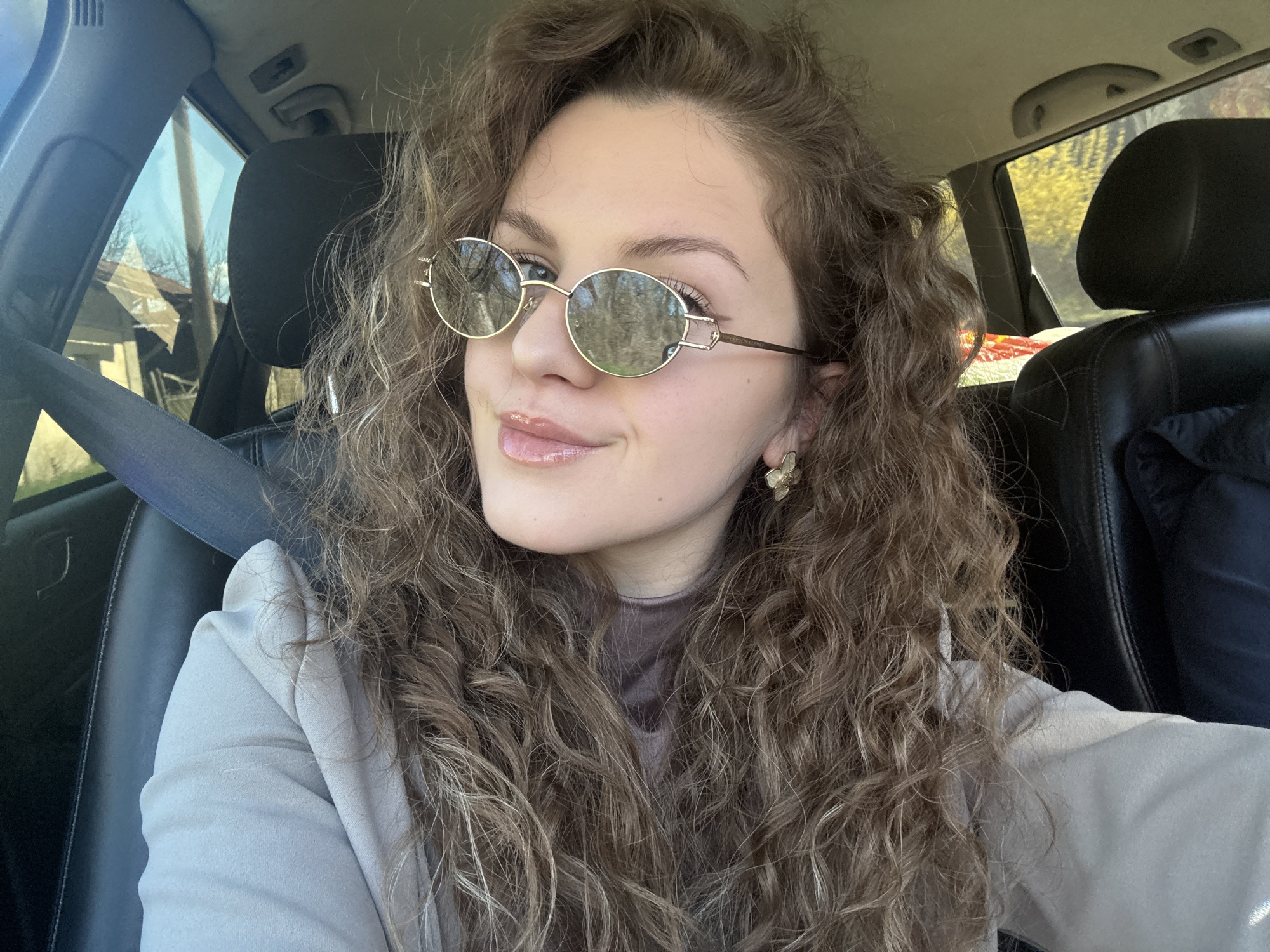 A young woman with curly hair and sunglasses is sitting in the driver's seat of a car. She is wearing a seatbelt, a beige blazer, and a turtleneck shirt. The car has black leather seats and the background shows foliage through the windows. The location is unknown, but not necessarily Nikolaevo, Bulgaria.