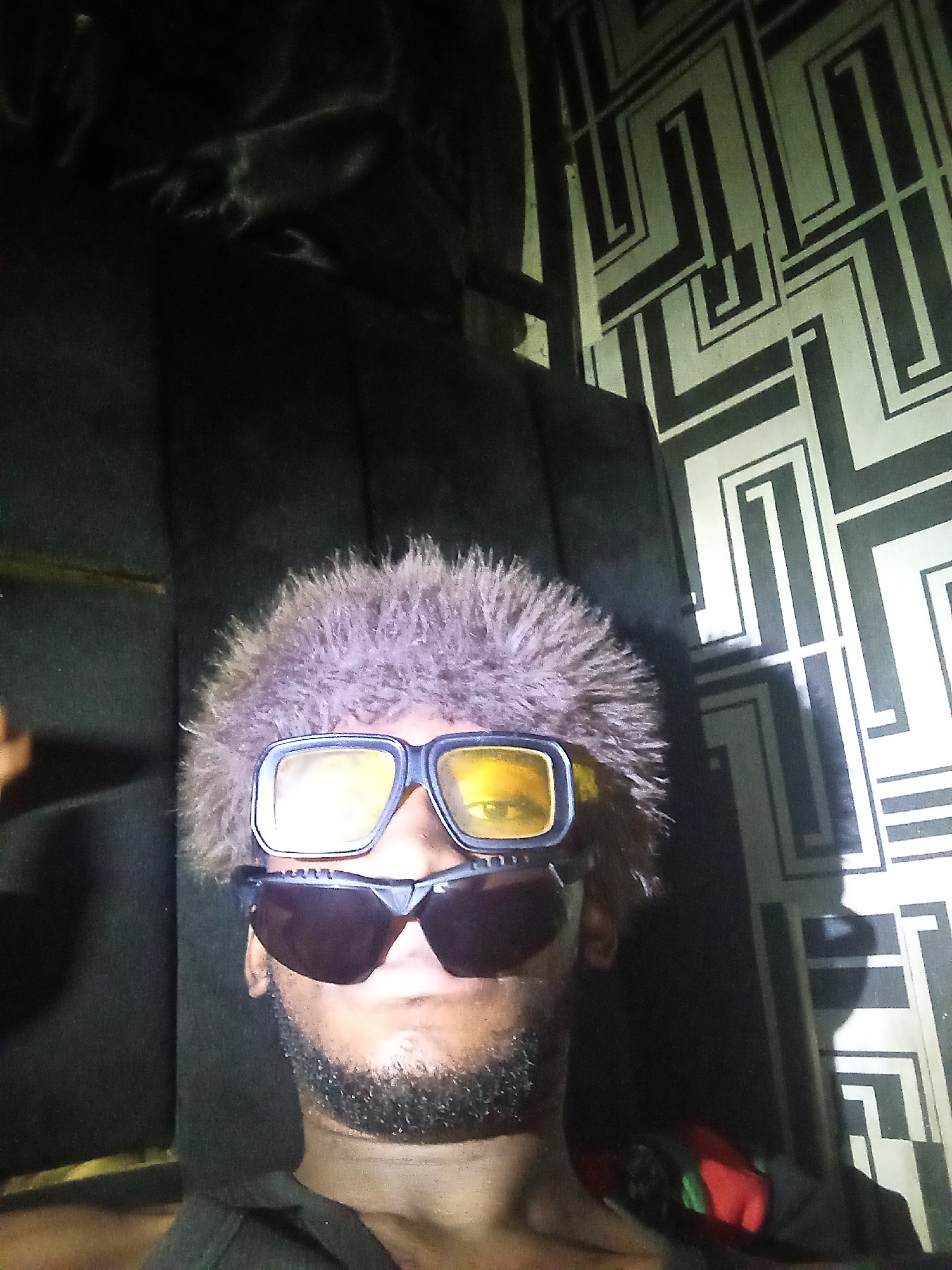 A selfie of a Black man wearing two pairs of glasses and a fluffy hat is taken indoors, possibly in Lokoja, Nigeria. The man has a beard and is looking directly at the camera. The background features a black, textured surface on the left and a wall with a geometric black and white pattern on the right. The lighting is dim.