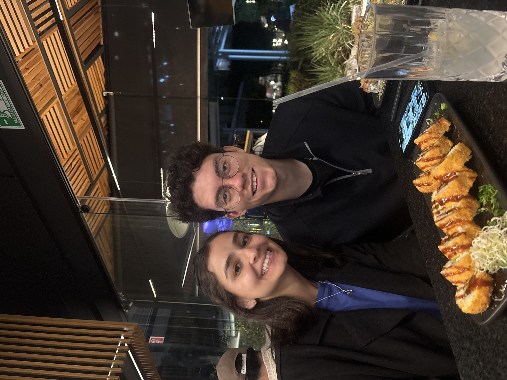 Two individuals are posing for a photograph at a restaurant. A man with curly dark hair and glasses is positioned behind a woman with dark hair. They both smile at the camera. A plate of fried sushi rolls is visible on the table in front of them, as well as a glass with a straw. The location is San Bernardino Tlaxcalancingo, Mexico.
