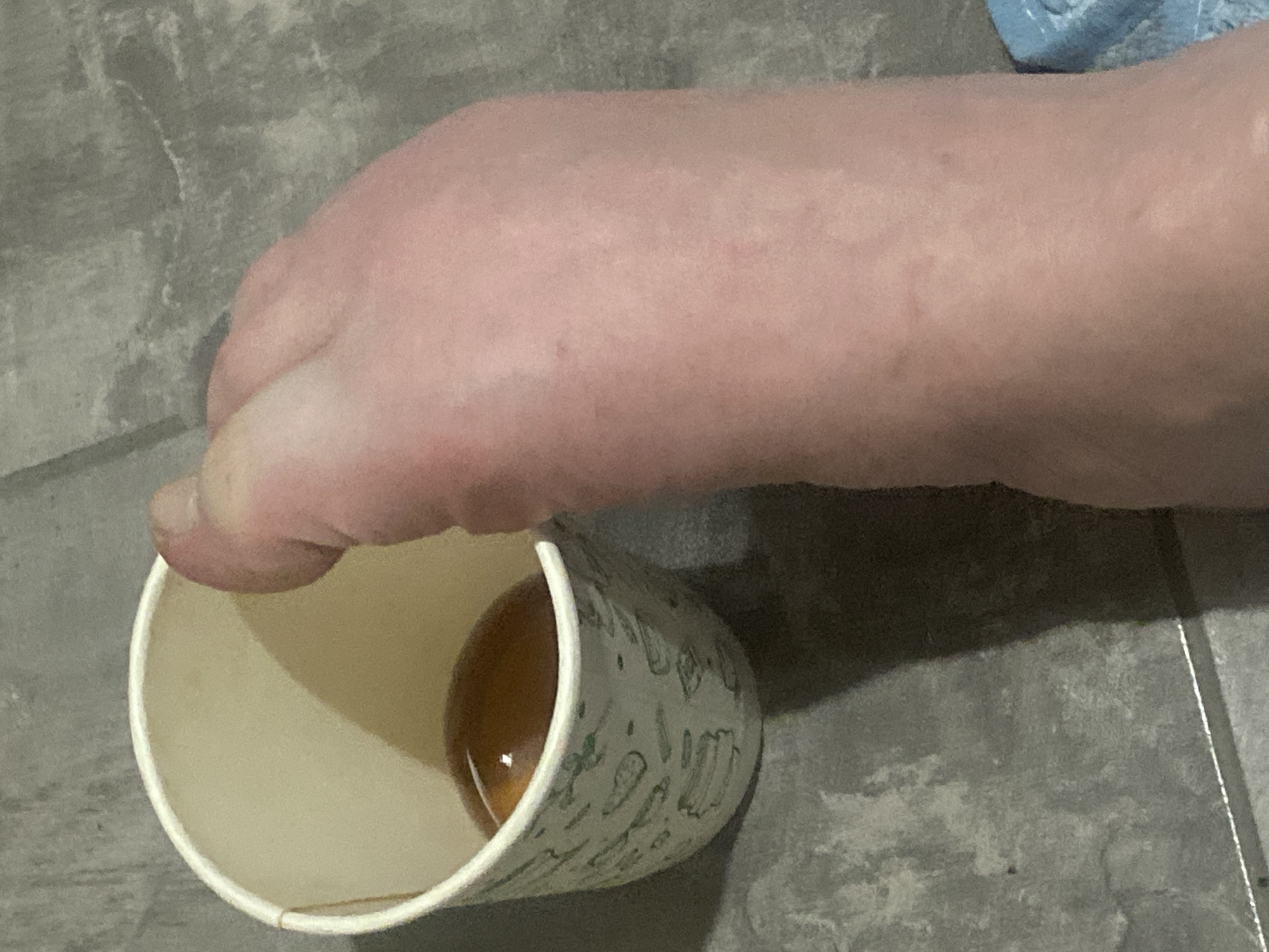 A close-up shot shows a person's foot holding a paper cup. The foot is positioned from the side, with the toes gripping the rim of the cup. The cup is light-colored with a subtle pattern of green designs. Inside the cup, a small amount of amber-colored liquid is visible. The background consists of a gray, textured surface, possibly concrete or tile flooring. A shadow from the foot and cup falls across the floor, suggesting indoor lighting. The scene is simple and focuses on the interaction between the foot and the cup. No text is visible in the image. The overall impression is a casual, everyday moment.