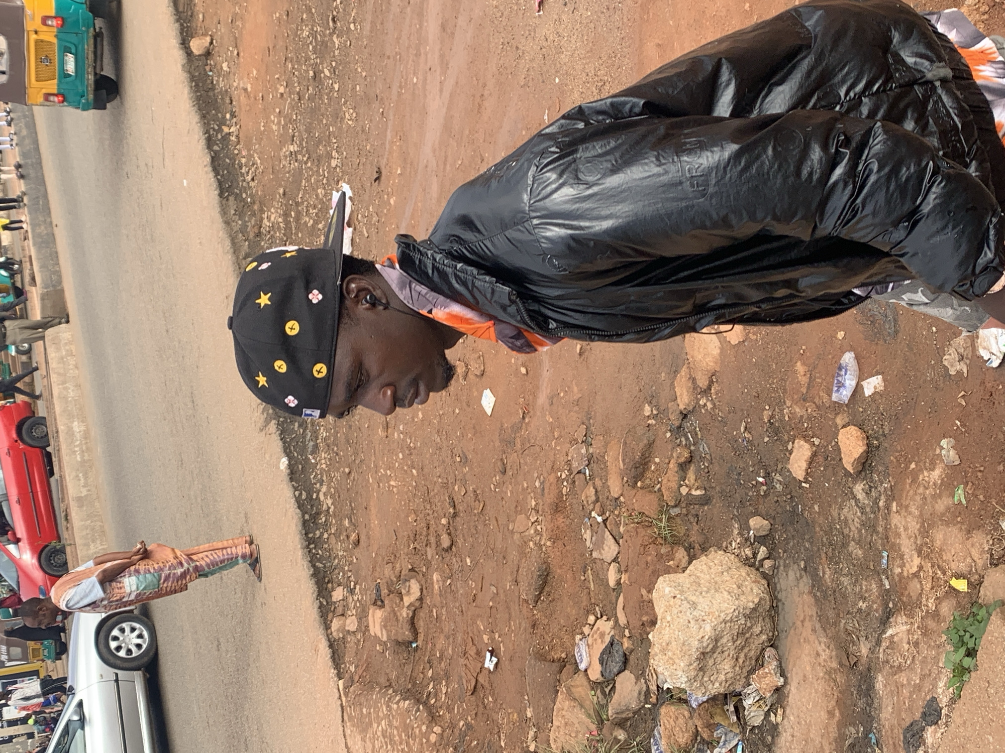 This image captures a street scene in Gura Top, Nigeria. A young Black man, wearing a black puffer jacket and a black baseball cap adorned with yellow stars and flowers, is the primary subject. He is looking to his right with a neutral expression. In the background, a red car is parked on the left side of the road, and further down, a yellow and green auto-rickshaw is visible. The ground is a mix of dirt and rocks, with some scattered litter. Another person, wearing a patterned garment, is also visible in the mid-ground on the left, near a silver car. The overall setting appears to be a roadside, suggesting a bustling urban or semi-urban environment. The lighting indicates it is daytime, and the presence of dirt and scattered items suggests a somewhat unkempt area. No visible text is discernible in the image.