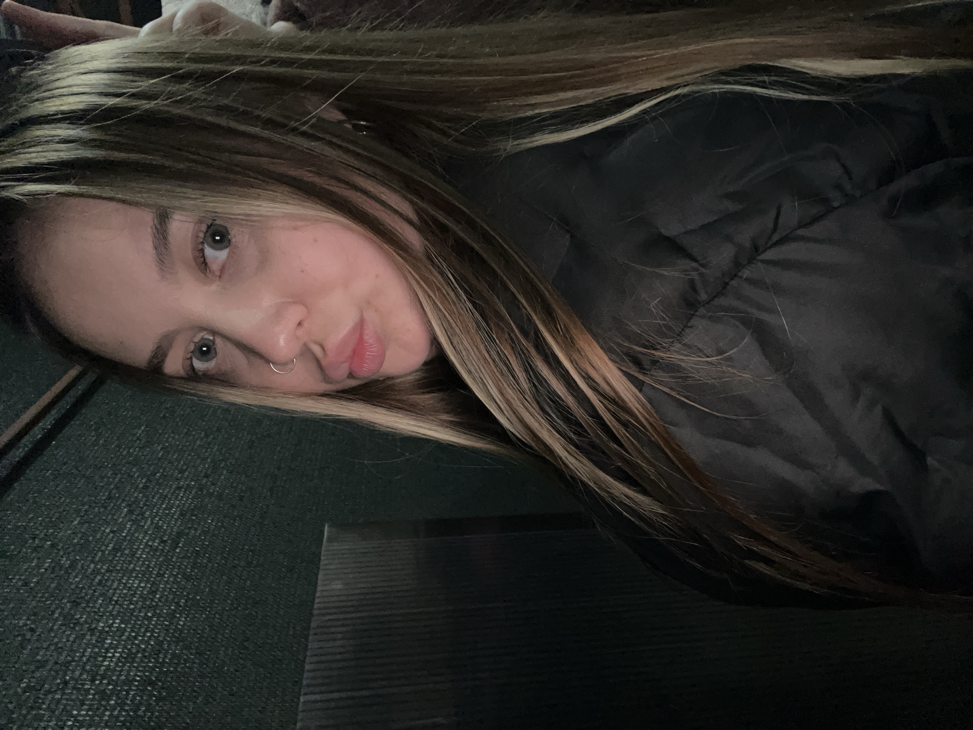 A woman with long brown hair is lying down, wearing a black puffer jacket. She has a nose ring and light eyes. The background is a dark, possibly green, textured surface. The image appears to be taken from a slightly elevated, angled perspective. The location is not possible to determine from the visual information provided.