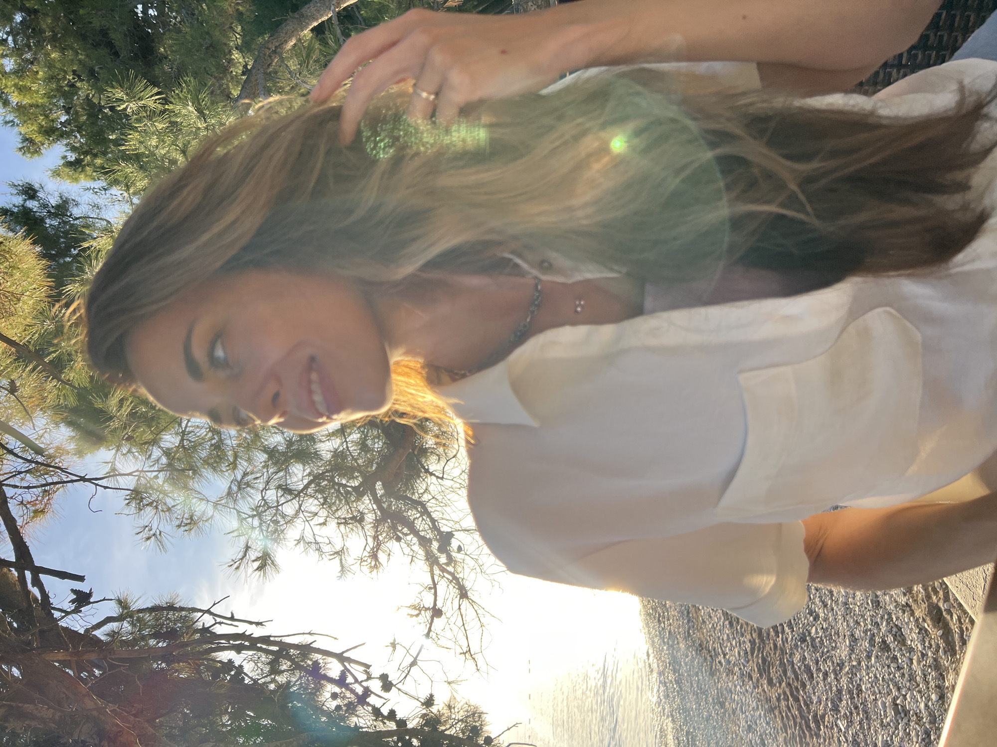 The image presents a vibrant, sun-drenched outdoor portrait of a young woman in Bar, Montenegro. The woman is positioned on the right side of the frame, captured from the chest up, looking towards the left with a soft, content smile. She has long, light brown hair, which appears to have blonde highlights, and her right hand is gently placed in her hair near her head, adorned with a ring. She is wearing a light-colored, possibly white, collared shirt and a subtle necklace.
The setting is characterized by strong, warm sunlight, indicating either early morning or late afternoon. The sun, peeking through the foliage, creates noticeable lens flares that shimmer across her hair and throughout the image. The background features dense, green coniferous trees, possibly pines, framing the top and bottom left sections of the photograph. Between the trees, a clear blue sky is visible. To the lower right, a calm body of water, likely the Adriatic Sea, stretches towards a pebbly beach or shoreline, intensely reflecting the golden sunlight. The overall impression is one of peaceful relaxation and natural beauty in a coastal environment. There is no visible text in the image.