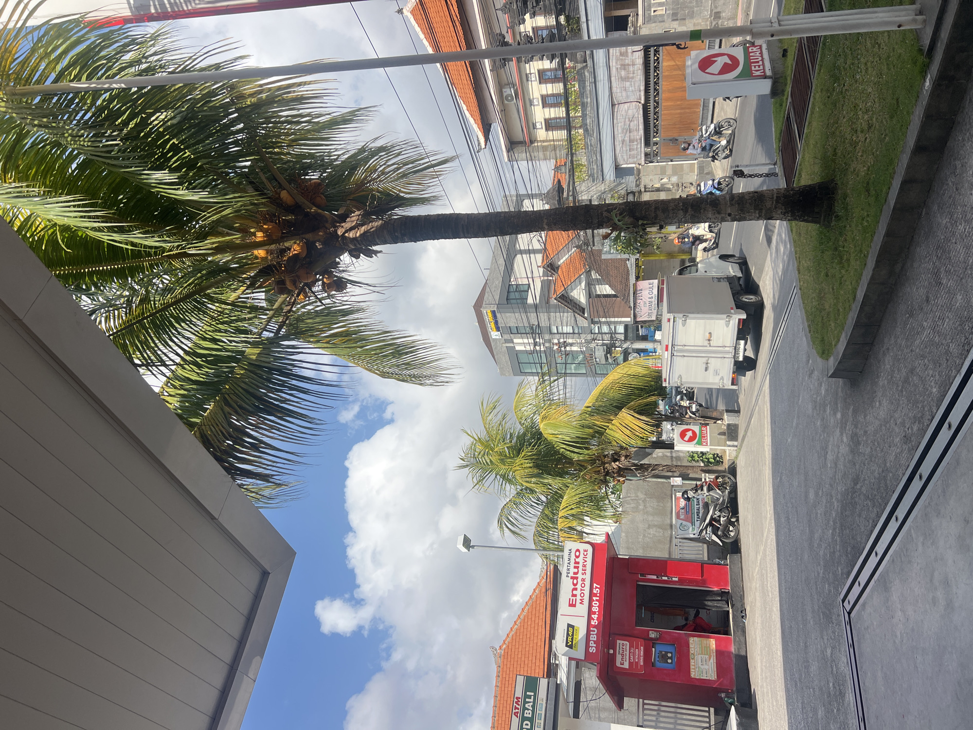 In Denpasar, Indonesia, a sunny day is captured with a palm tree laden with coconuts in the foreground. To the left, a red structure with "Pertamina Enduro Motor Service" signage and "SPBU 54.801.57" is visible, likely a gas station booth. A white truck is driving away from the camera, and several motorcycles are on the road. A sign with a red arrow and the word "KELUAR" (exit) in white lettering is on the right side of the road. Buildings with orange tiled roofs and a modern grey building are in the background. The sky is blue with scattered clouds.