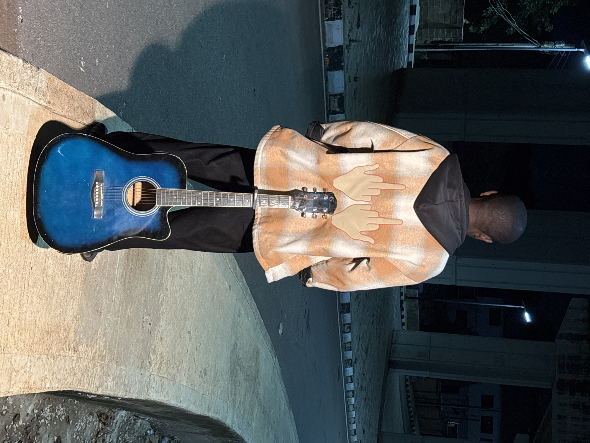 The image shows a person standing with their back to the camera, holding an acoustic guitar. The person is wearing a plaid shirt with cartoon hands giving the middle finger printed on the back, and black pants. The guitar is blue and resting on the ground. The scene appears to be outdoors at night, with streetlights and large concrete pillars visible. The ground is paved with asphalt and concrete. Based on the architectural style of the concrete pillars and the general urban aesthetic, the location is consistent with areas in Jos, Nigeria, where elevated roadways and underpasses are common, often illuminated by streetlights for safety and navigation.
