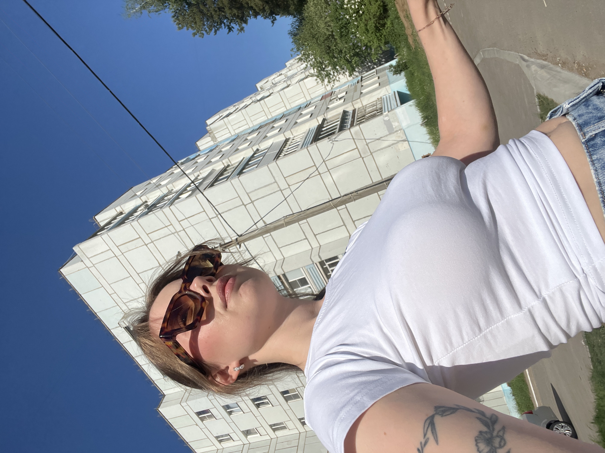 A low-angle photograph shows a woman wearing sunglasses and a white T-shirt, with a large, concrete apartment building in the background. Clear blue sky is visible above the building. The setting is likely in Chaikovsky, Russia, indicated by the architectural style of the apartment block. A narrow road and greenery are partially visible in the foreground. The woman’s arm is raised, and she has a tattoo on her inner arm. The photograph appears to be taken at midday.
