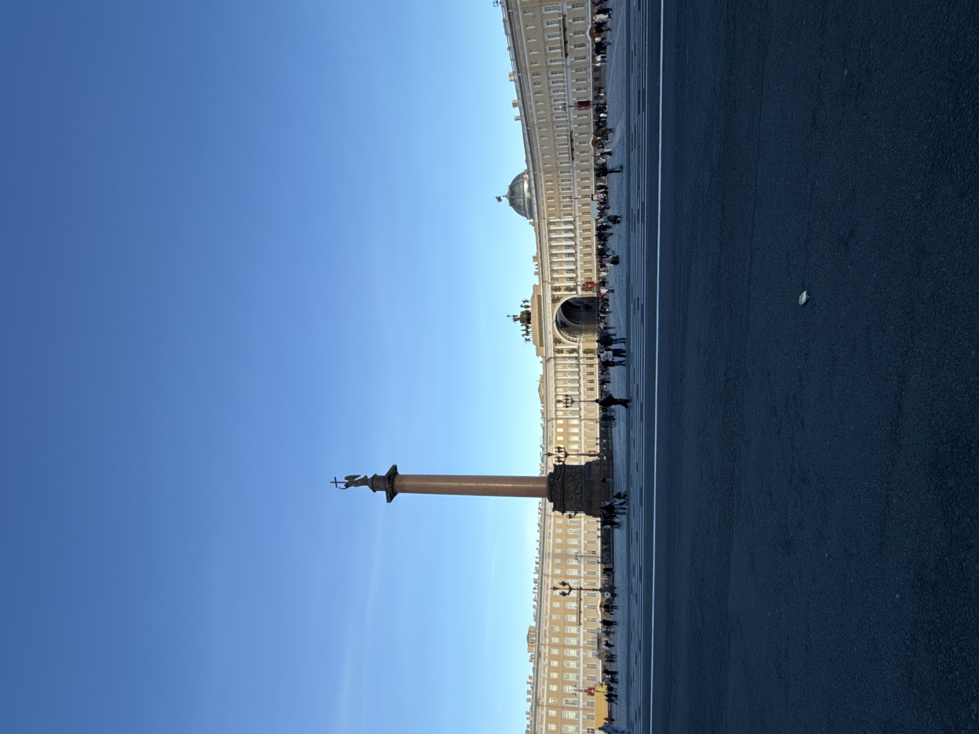 This image captures a vast, sunlit public square, specifically Palace Square in Saint Petersburg, Russia. The scene is dominated by the towering Alexander Column, positioned prominently in the mid-ground. The column is a reddish-brown monolithic structure, surmounted by a statue of an angel holding a cross.
To the right, a long, curved neoclassical building, identified as the General Staff Building, stretches across the background. This ornate structure features numerous windows, columns, and architectural details. A large dome, likely part of the nearby Winter Palace or Hermitage Museum, is partially visible above the General Staff Building in the distance.
The square itself is paved with dark asphalt or stone, traversed by distinct white lines that suggest pedestrian or traffic markings. Numerous people are scattered across the square and along the base of the General Staff Building. They appear to be pedestrians or tourists, casually walking and observing the historic surroundings. The activity suggests a typical day of public access and sightseeing at a major landmark.
The weather is clear and bright, with a vibrant, cloudless blue sky filling the upper portion of the frame, indicating a sunny day, likely morning or early afternoon. The overall atmosphere is peaceful and calm. No visible text is discernible in the image.