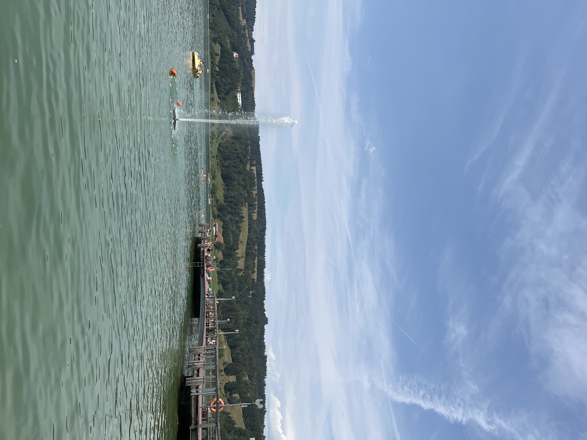 The image depicts a scenic lake with a pier and several recreational activities. The lake is a vibrant green, with gentle ripples on its surface. In the foreground, the water is clear, with a few small specks visible. A fountain sprays water high into the air in the middle of the lake, creating a dynamic visual element.
To the left of the fountain, a yellow paddle boat with two people is visible. Further out in the lake, other small boats and swimmers can be seen enjoying the water. A long wooden pier extends into the lake from the right side of the image. Several people are seated and standing on the pier, some appearing to be relaxing or observing the water. A life preserver ring hangs on a post on the pier, indicating a safety measure.
The background features a lush, green hillside covered in dense forest, dotted with some buildings. The sky above is a clear blue, with wispy white clouds streaking across it.
This scene is located at the Kleiner Alpsee in Immenstadt im Allgäu, Germany. The Kleiner Alpsee is a popular recreational area, known for its clear waters and surrounding natural beauty, offering opportunities for swimming, boating, and enjoying the outdoors. The presence of the pier, fountain, and various boats suggests a well-established public access point for leisure activities.