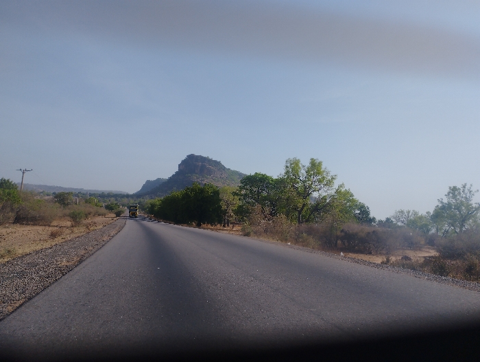 The image shows a paved road stretching into the distance, with a single vehicle traveling toward the viewer. On either side of the road are trees and shrubs. In the background, there is a large, rocky hill. The sky is a light blue with some haze. The image was taken in the city of Ture-Balam, Nigeria, at the coordinates 9.807 latitude, 11.381 longitude.