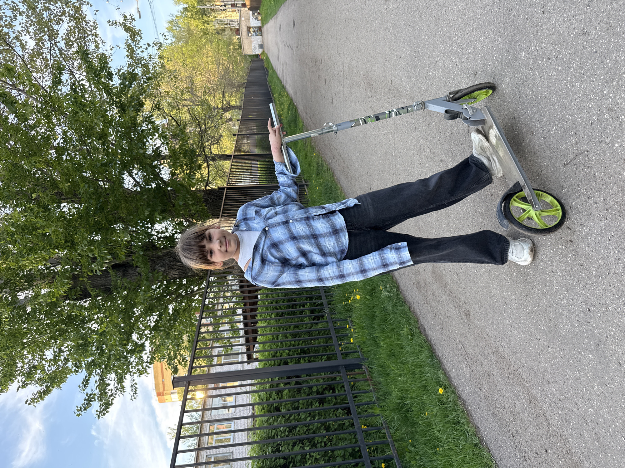 A youth is standing on a kick scooter on a paved path adjacent to a grassy area. The person wears black jeans, white sneakers, and a blue plaid shirt over a white undershirt. They are holding a mobile phone in their left hand. A black metal fence runs along the edge of the path, and behind the fence are buildings, partially obscured by trees. The background shows trees and buildings under a slightly overcast sky. The location is likely Rusanovo, Russia, based on the visible architecture.