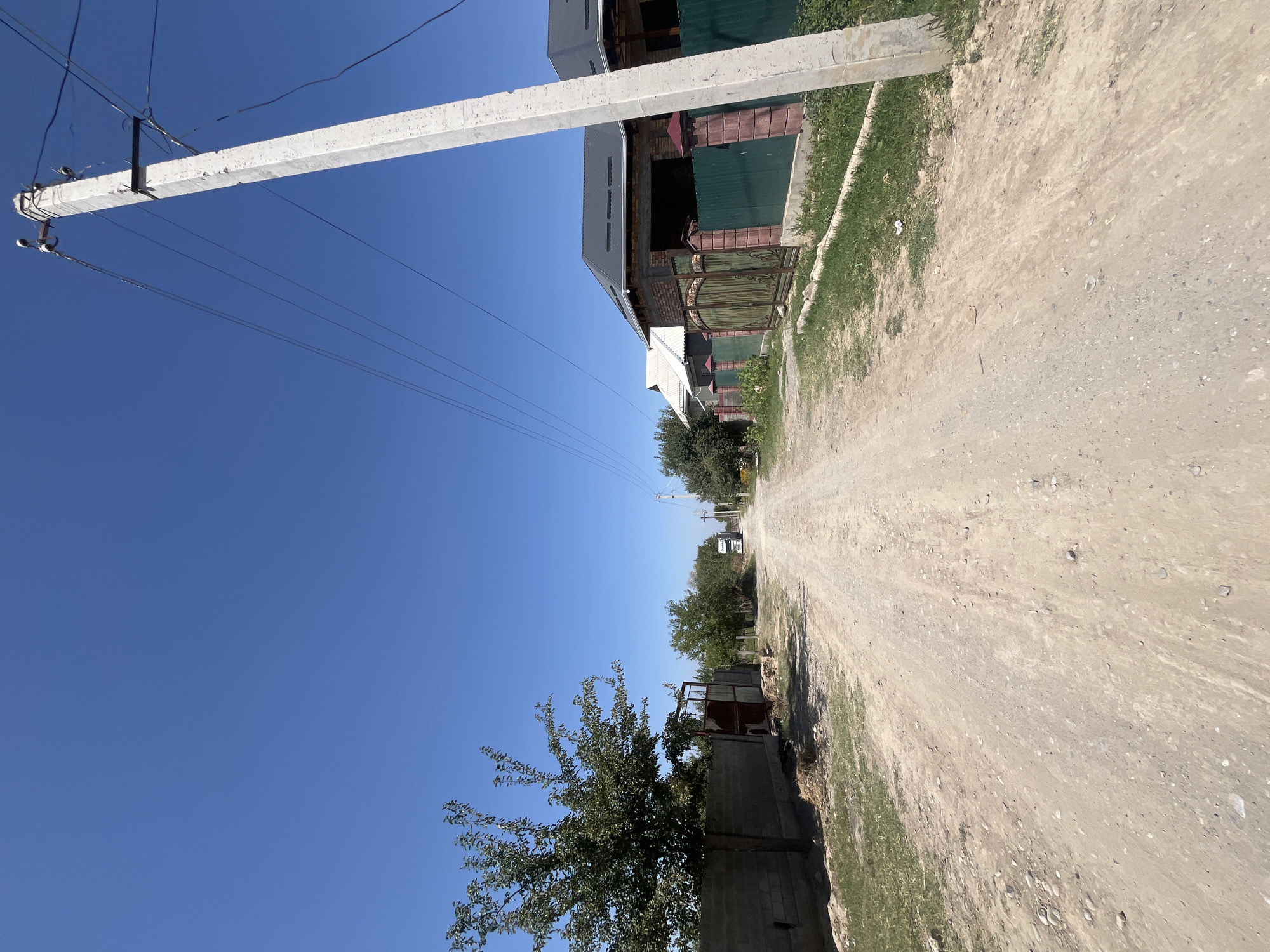 The image shows a rural road in clear daylight under a bright blue sky. The road is unpaved, with visible dirt and small stones. On the left side of the road, a low concrete wall is visible, with a tree growing behind it. Further down the road, a rusty metal gate is present. On the right side of the road, a power pole stands prominently, with several electrical wires extending from it. Beyond the pole, a fence made of green metal panels and brick pillars separates the road from a property with a house featuring a dark grey roof. The road extends into the distance, where a white truck can be seen. The environment appears to be a residential area with sparse vegetation.