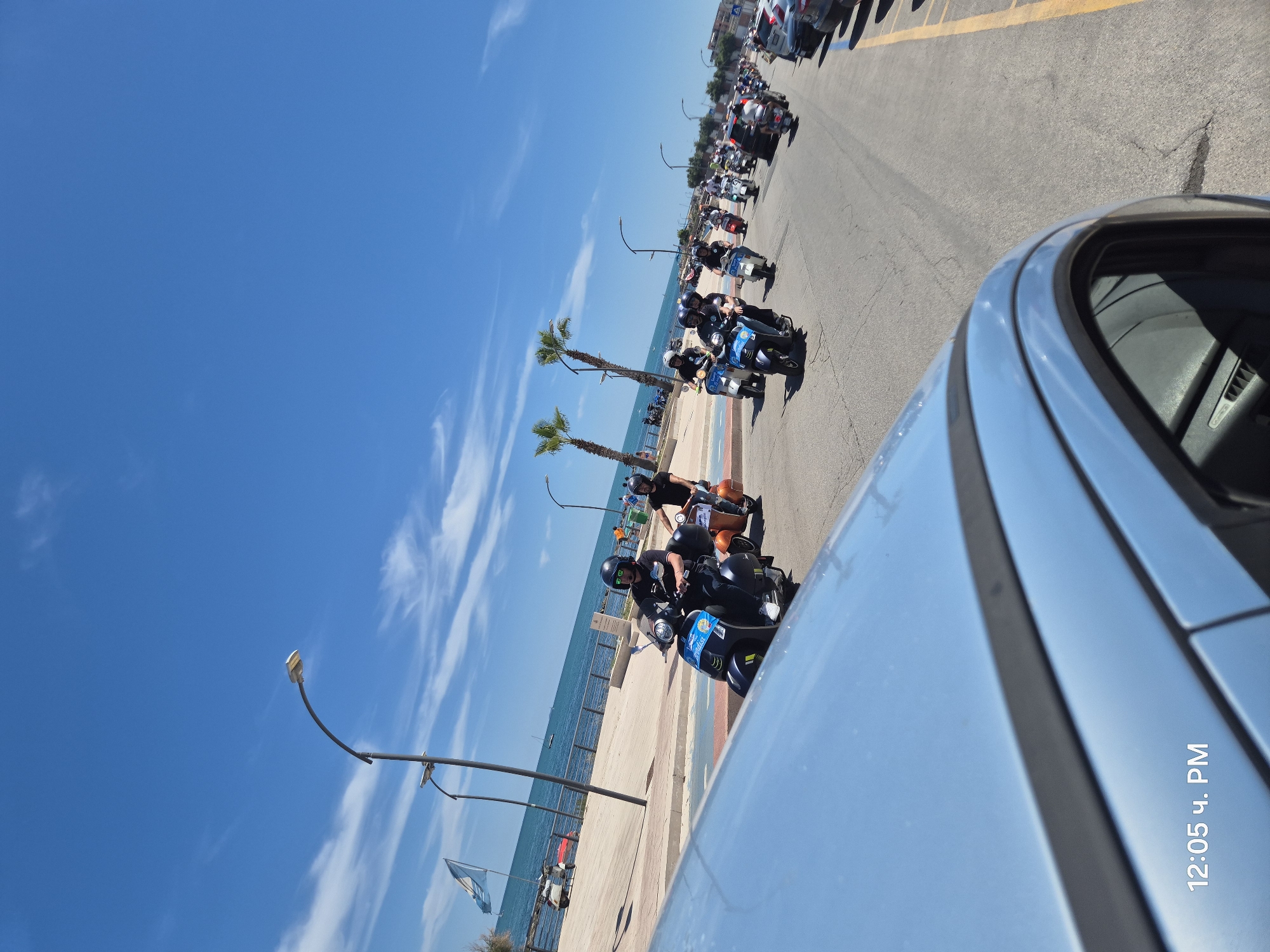 The image shows a motorcade of Vespas traveling along a coastal road. The sky is clear blue with scattered white clouds. The road is paved and bordered by a sidewalk and a body of water, likely the Adriatic Sea. On the sidewalk, there are palm trees, streetlights, and a flag. The Vespas are mostly black and white, with a few orange ones. The riders are wearing helmets and casual clothing. The perspective is from a vehicle moving alongside the Vespas, with the hood of the vehicle visible in the foreground. The timestamp "12:05 ч. PM" is visible in the lower left corner. This scene appears to be in Bisceglie, Italy, given the presence of coastal scenery and the Italian flag.