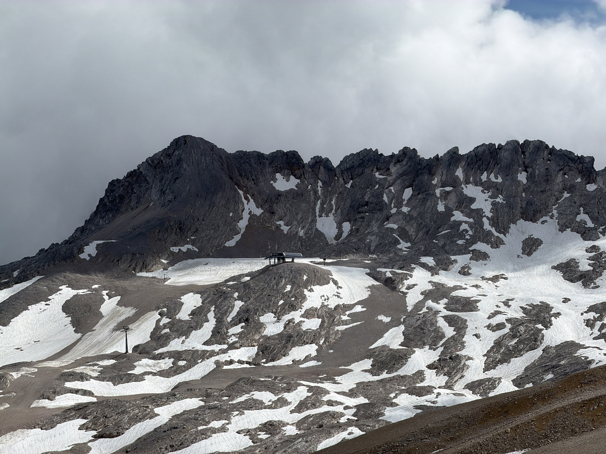 This is a photograph of the Zugspitze, the highest peak in Germany, located near Garmisch-Partenkirchen. The image captures a rugged, rocky mountain landscape partially covered in snow. A cable car station is visible on a rounded summit in the mid-ground, with its associated infrastructure, including cables and a support tower, also present. The sky is overcast with dramatic clouds, casting shadows on the mountainside. The terrain is a mix of scree, rock, and patches of snow, indicative of a high-altitude environment. The scene is devoid of any people.