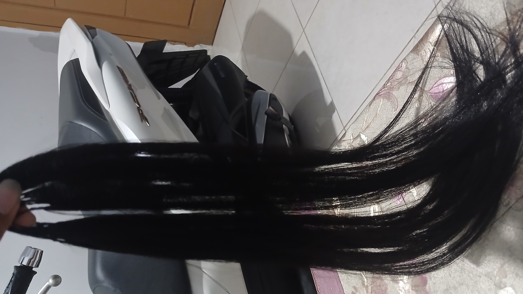 In Sampang, Indonesia, a close-up shot shows a person's hand holding several bundles of long, black hair. The hair hangs down, partially obscuring the background, which features a white and black scooter. The scooter has a distinctive design with silver accents and appears to be parked indoors on a tiled floor. A wooden door is visible in the far right of the frame. The lighting is dim, casting shadows and creating a slightly hazy atmosphere.