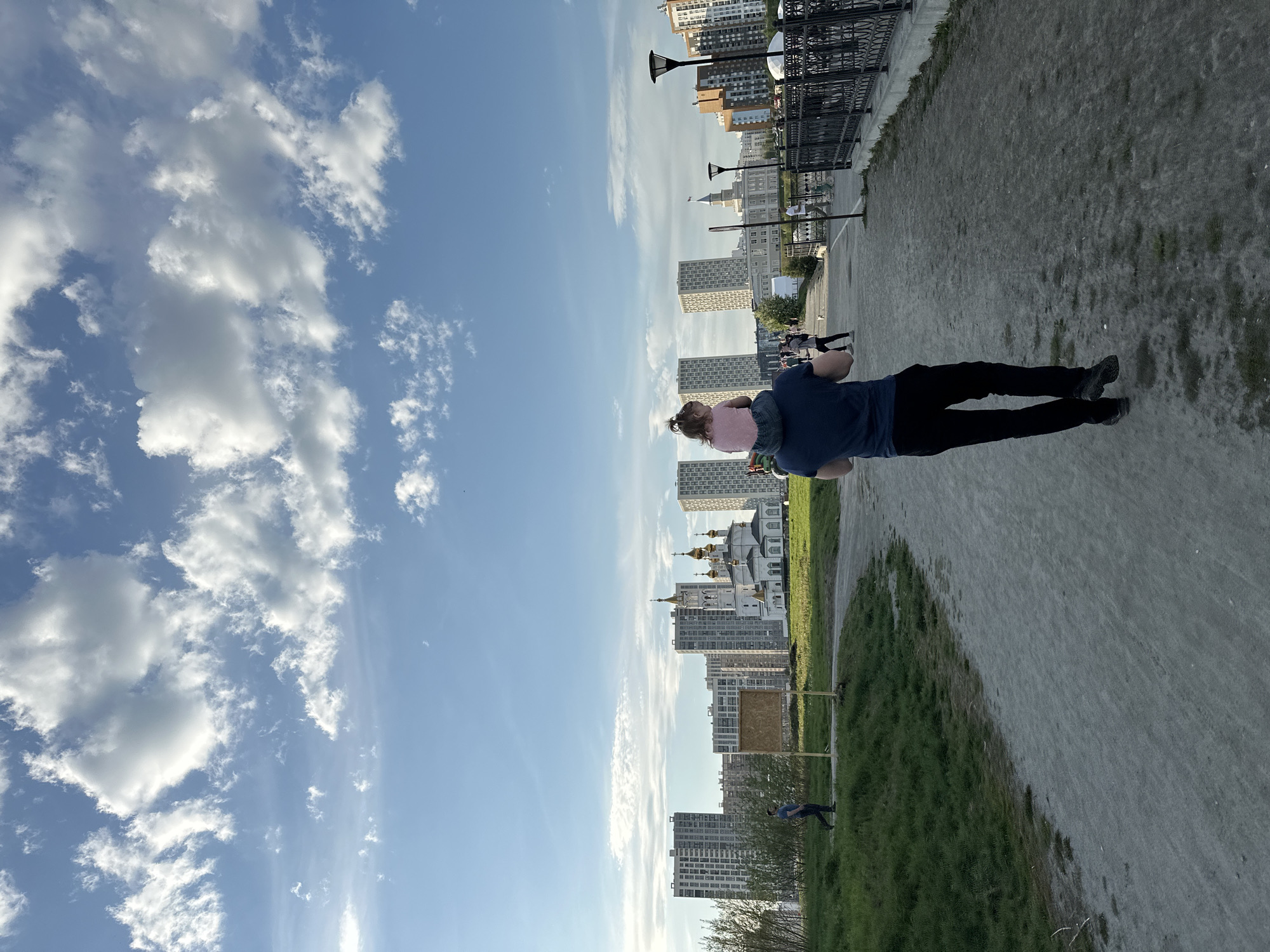 A man carries a child on his shoulders while walking in a park in Yekaterinburg, Russia. The man is wearing a dark blue shirt and dark pants. The child is wearing a pink shirt. Behind them, there are buildings, including a church with gold domes. The sky is blue with white clouds. The park has a path and grassy areas. There is a metal railing in the background.