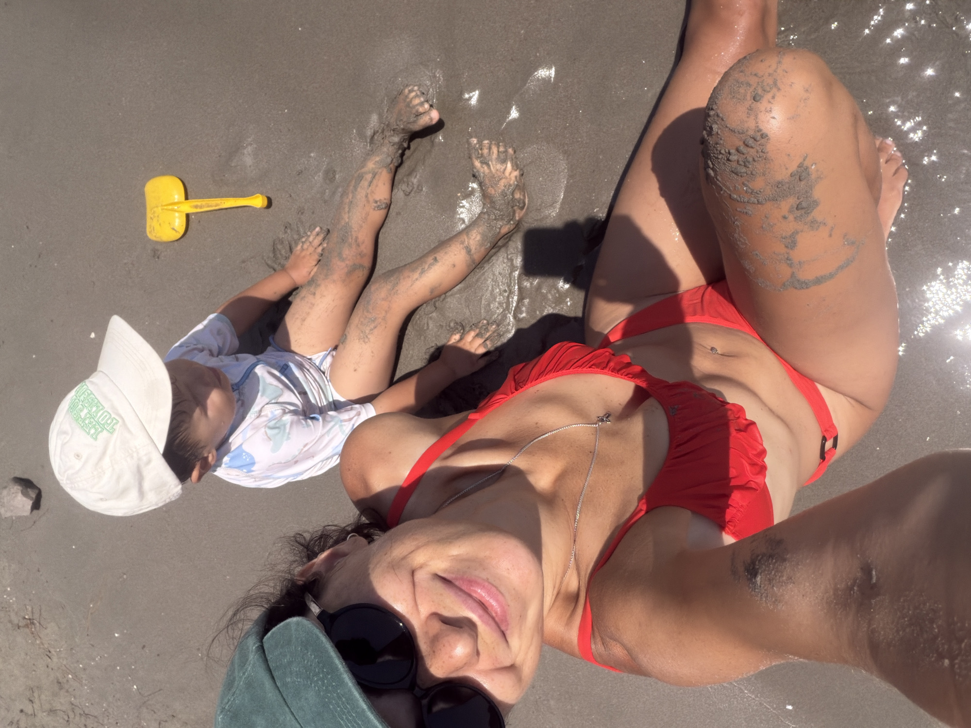 In Bigova, Montenegro, two individuals are on a sandy beach. In the foreground, a woman wearing a red bikini and a green cap is taking a selfie. She has sunglasses on and is smiling at the camera. To her right, a child wearing a white baseball cap and a blue and white patterned swimsuit sits on the sand, covered in sand. A yellow plastic shovel lies on the sand between the woman and the child. The environment is a beach with wet sand and some glistening water.