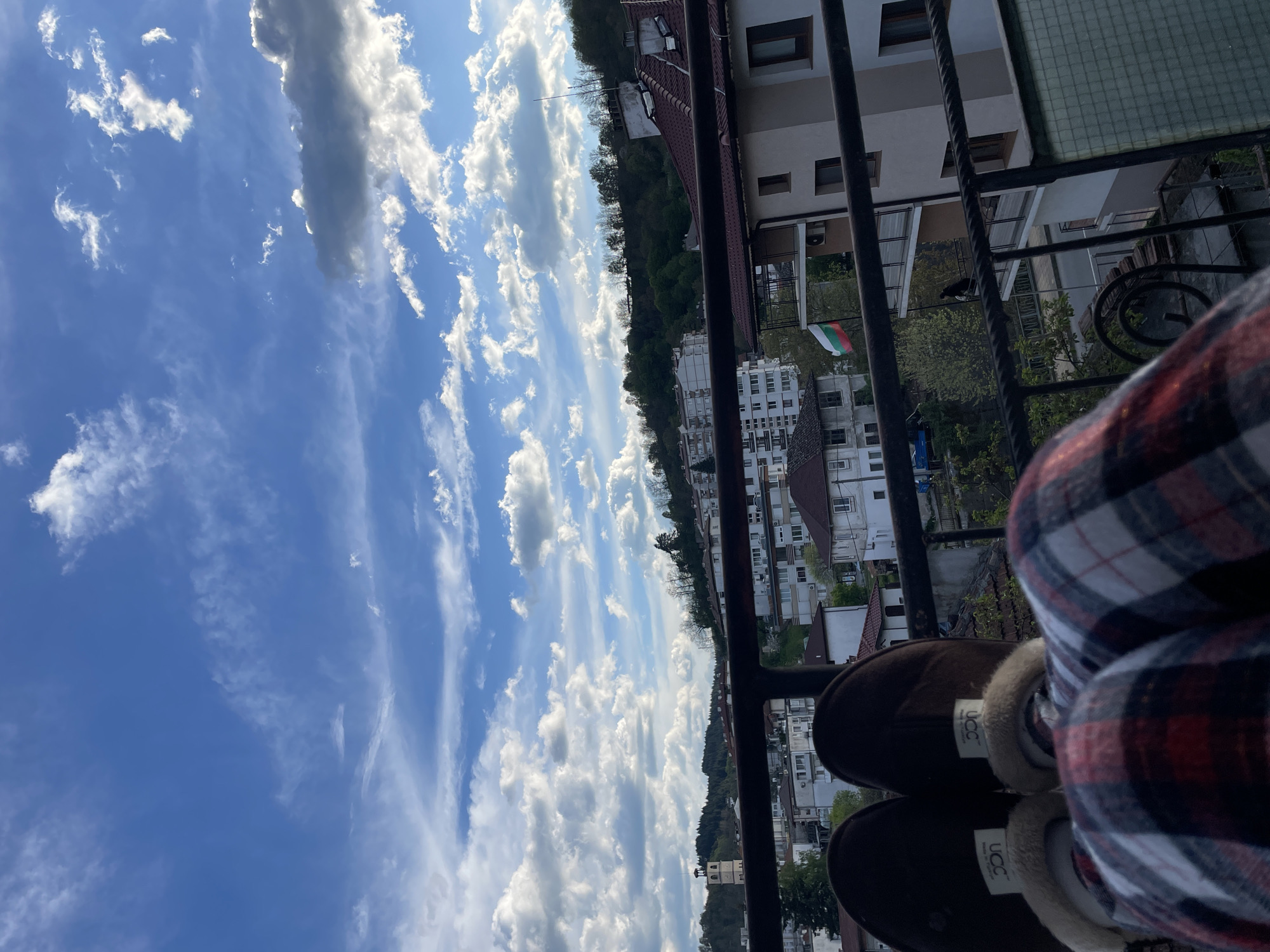 The image shows a view from a balcony in Zlatograd, Bulgaria. The sky is predominantly blue with scattered white clouds. In the mid-ground, there are residential buildings and trees. A Bulgarian flag is visible hanging from a balcony. A metal railing is in the foreground, partially obscuring the view. In the lower right of the image, a person is sitting with their legs crossed, wearing plaid pants and UGG brand slippers.