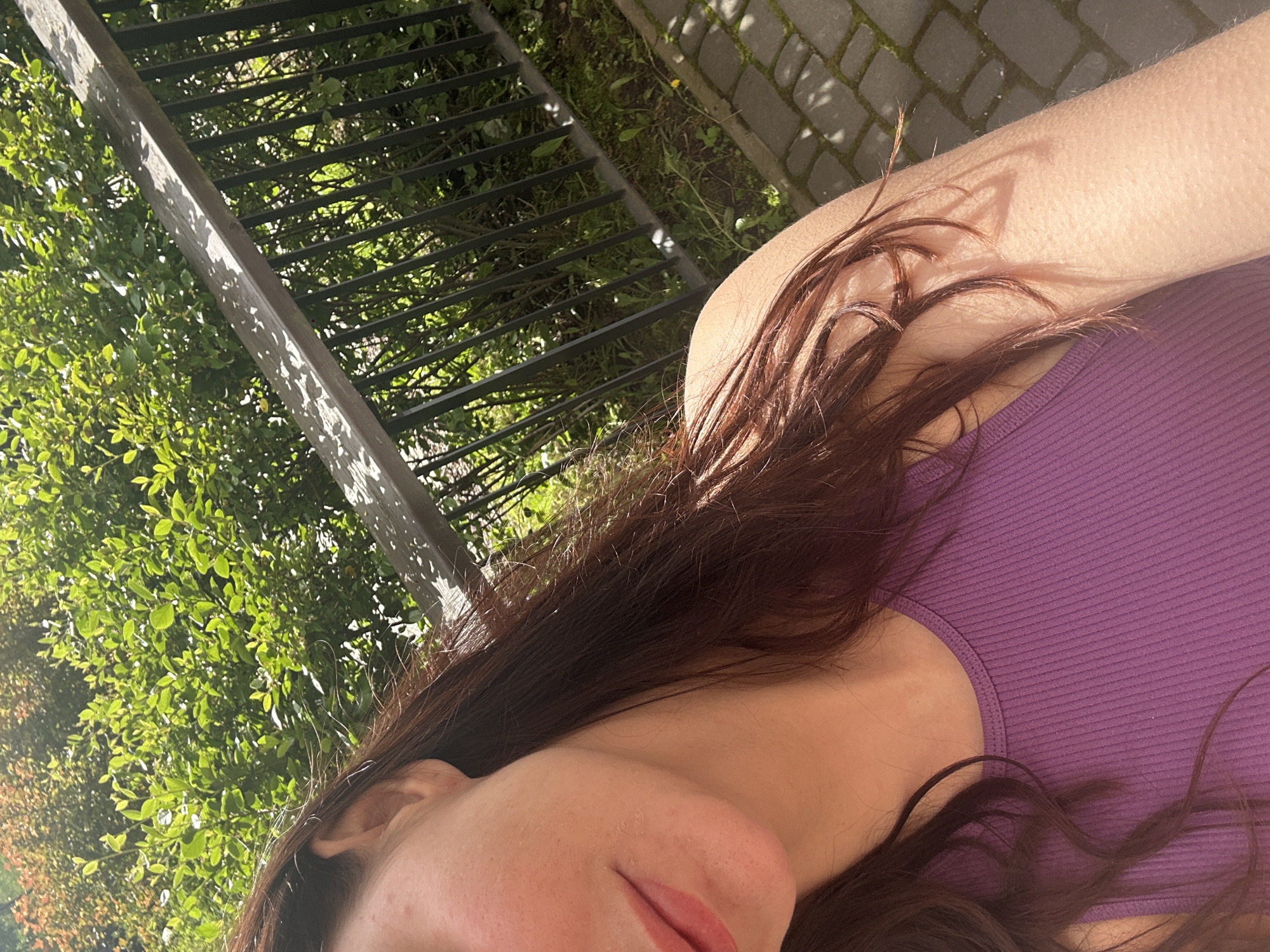 A young woman is shown from the chest up, outdoors, with lush green foliage behind her. She is wearing a purple tank top. Sunlight illuminates her skin, creating a dappled shadow effect from the railing visible in the background. The ground appears to be paved with bricks or stone. Her long, dark brown hair is visible, framing her face and shoulders. The image is likely taken during the daytime in a garden or park setting.