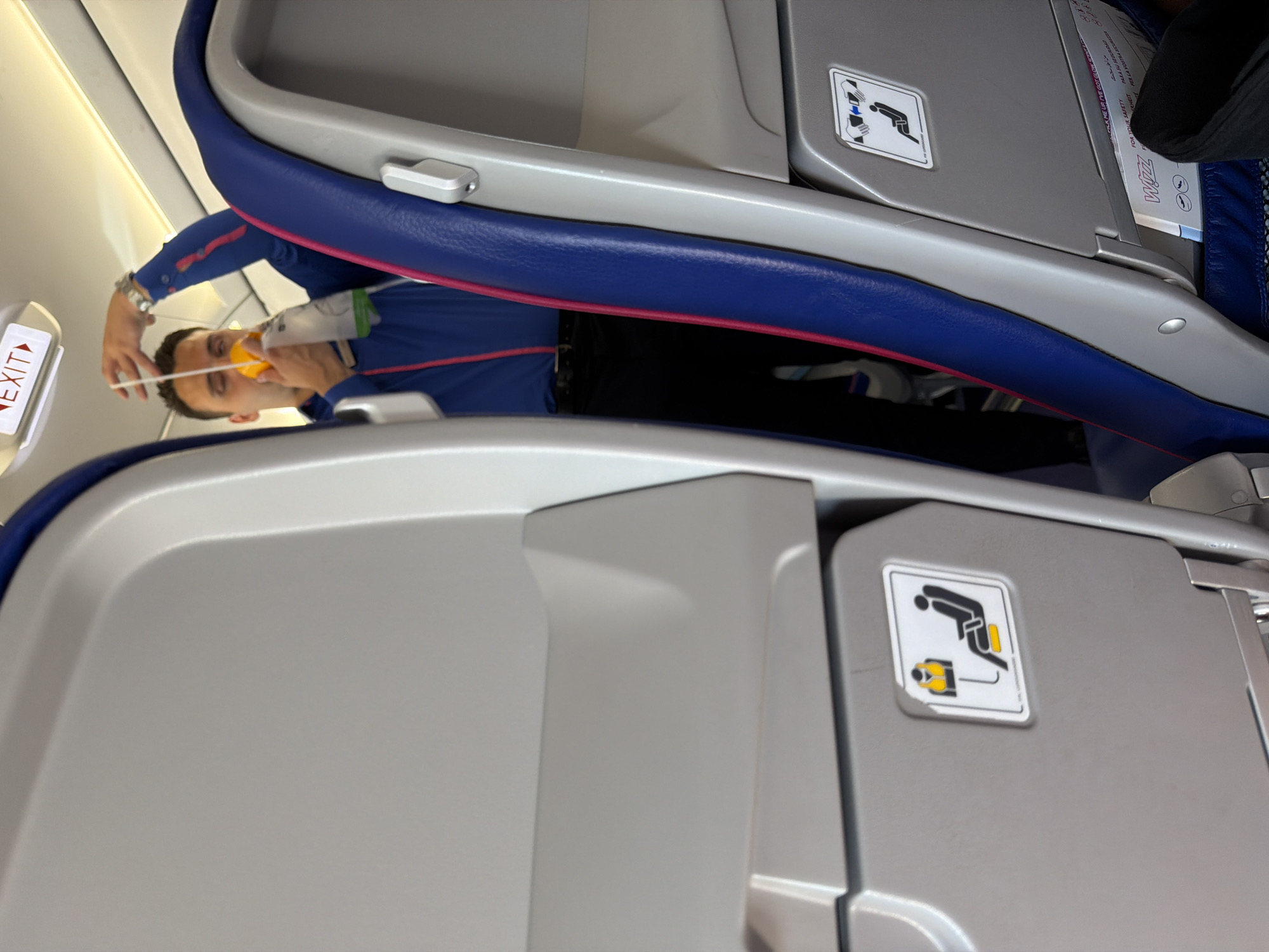 A man in a blue shirt and black pants is holding an oxygen mask in front of his face inside an airplane cabin. He appears to be demonstrating how to use the mask, with the mask hanging from a white string. The cabin interior is visible, with gray seats and blue and pink accents on the seatbacks. An "EXIT" sign is illuminated above the man. Below the man's left hand is a safety card with an icon of a person in a life vest. Another safety card is visible on the seat in front of him. The image is a medium shot, taken from a slightly low angle. The scene is forensic, matter-of-fact, and describes the physical elements of the image without interpretation. The location of Donja Cijevna, Montenegro, is not depicted or suggested by the image.