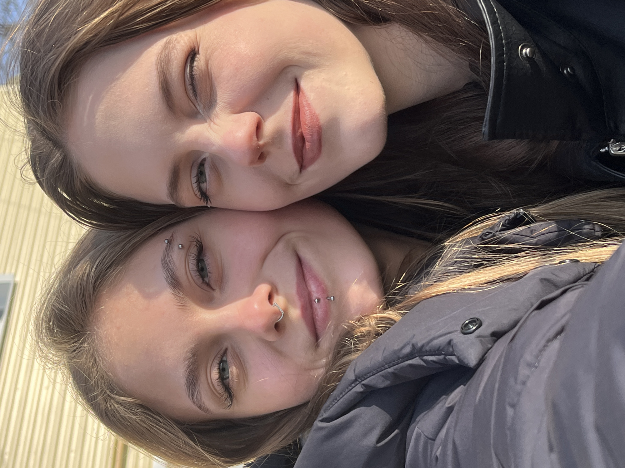 Two young women are captured in a close-up selfie, embracing a casual and friendly pose in an outdoor setting in Фоки, Russia. The image is rotated, with their heads angled to the bottom left.
The woman positioned higher in the frame has light brown hair, which appears to have lighter streaks, and is wearing a black, leather-like jacket. She sports subtle makeup, including mascara and a natural-toned lipstick, and has a small nose piercing. Her face is lightly tanned, and she offers a gentle, closed-mouth smile.
Below her, and slightly to the front, is the second woman, also with light brown, possibly blonde-streaked hair. She is wearing a dark grey or black padded jacket. This woman has several visible facial piercings: a vertical barbell piercing above her left eyebrow, a silver septum ring, and a philtrum (Medusa) piercing. She also has mascara applied and a subtle, friendly smirk.
Both women are looking directly at the camera, their faces illuminated by bright, natural sunlight, suggesting a clear day. The background behind them is a light-colored, possibly pale yellow or beige, building with vertical siding, indicating an outdoor, possibly residential, environment. No discernible text is visible in the image. The overall mood conveyed is one of youthful companionship and ease.