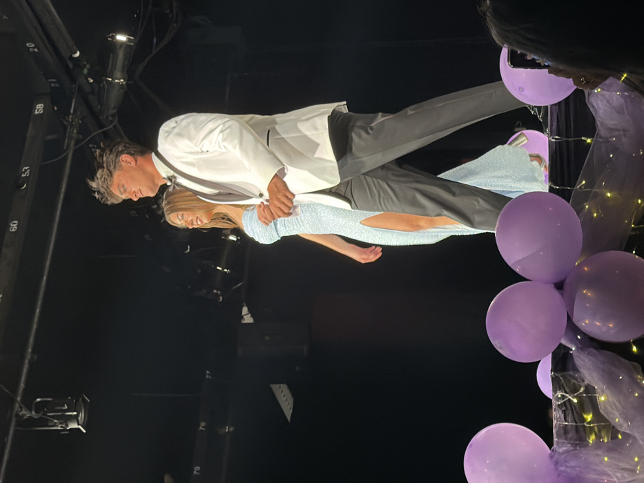 Two young individuals are depicted walking from left to right on a stage at a formal event in Palos Verdes Estates, United States. The male, positioned on the right, wears a white tuxedo jacket with black lapels, a black bow tie, a white shirt, and dark trousers. His hair is dark and styled upwards. The female, on the left, wears a light blue, strapless, shimmering floor-length gown featuring a high slit on the left side. She has long blonde hair. Both individuals are looking slightly upwards and to their right, exhibiting subtle smiles, and appear to be holding hands. The stage's lower foreground is decorated with multiple purple balloons and dark fabric draped with small yellow string lights. The background is dark, revealing overhead stage lighting rigs and metal trusses marked with white numbers such as "60," "53," and "20." A partial view of a person holding a smartphone with a purple case is visible in the bottom right corner, indicating an audience member.