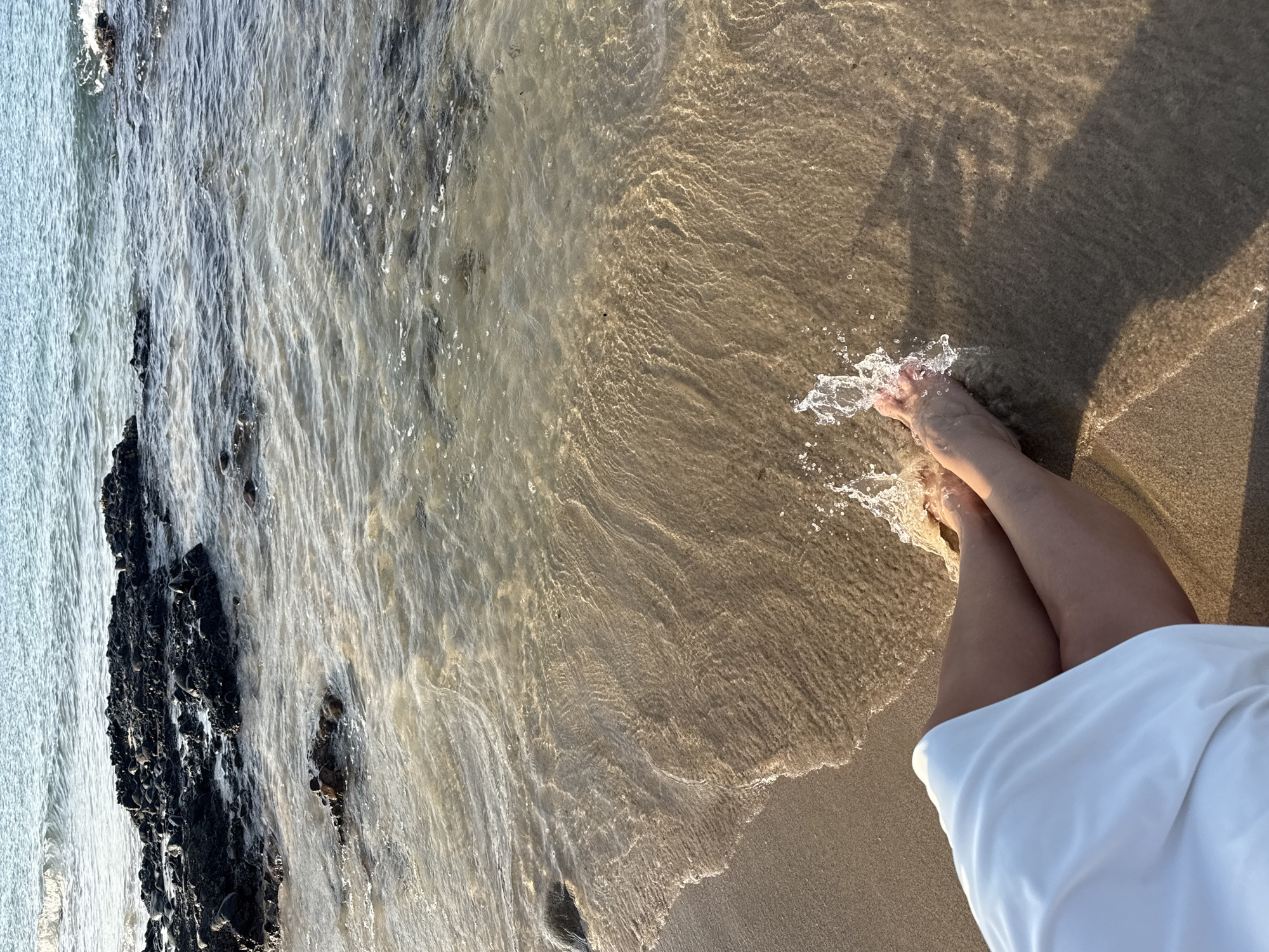 The image captures a first-person perspective of a person's legs and feet at the edge of the sea in Al Fiqait, United Arab Emirates. The person is standing in shallow, clear water that gently washes over their feet, creating splashes. They are wearing a white garment, possibly a dress or skirt, which is visible at the bottom right of the frame.
The scene is a tranquil beach setting, with golden-hued sand meeting the clear, rippling water. The water reveals the textured sandy bottom beneath. To the left, a dark, rocky shoreline or outcrop extends into the water, contrasting with the lighter tones of the sand and water. Further out, the water appears deeper and a lighter blue-grey.
The lighting suggests late afternoon or early morning, characterized by warm, golden tones on the sand and water, and the presence of elongated shadows, including the shadow of the person's upper body stretching onto the wet sand. The atmosphere appears calm and peaceful, inviting a sense of relaxation. No visible text is present in the image.