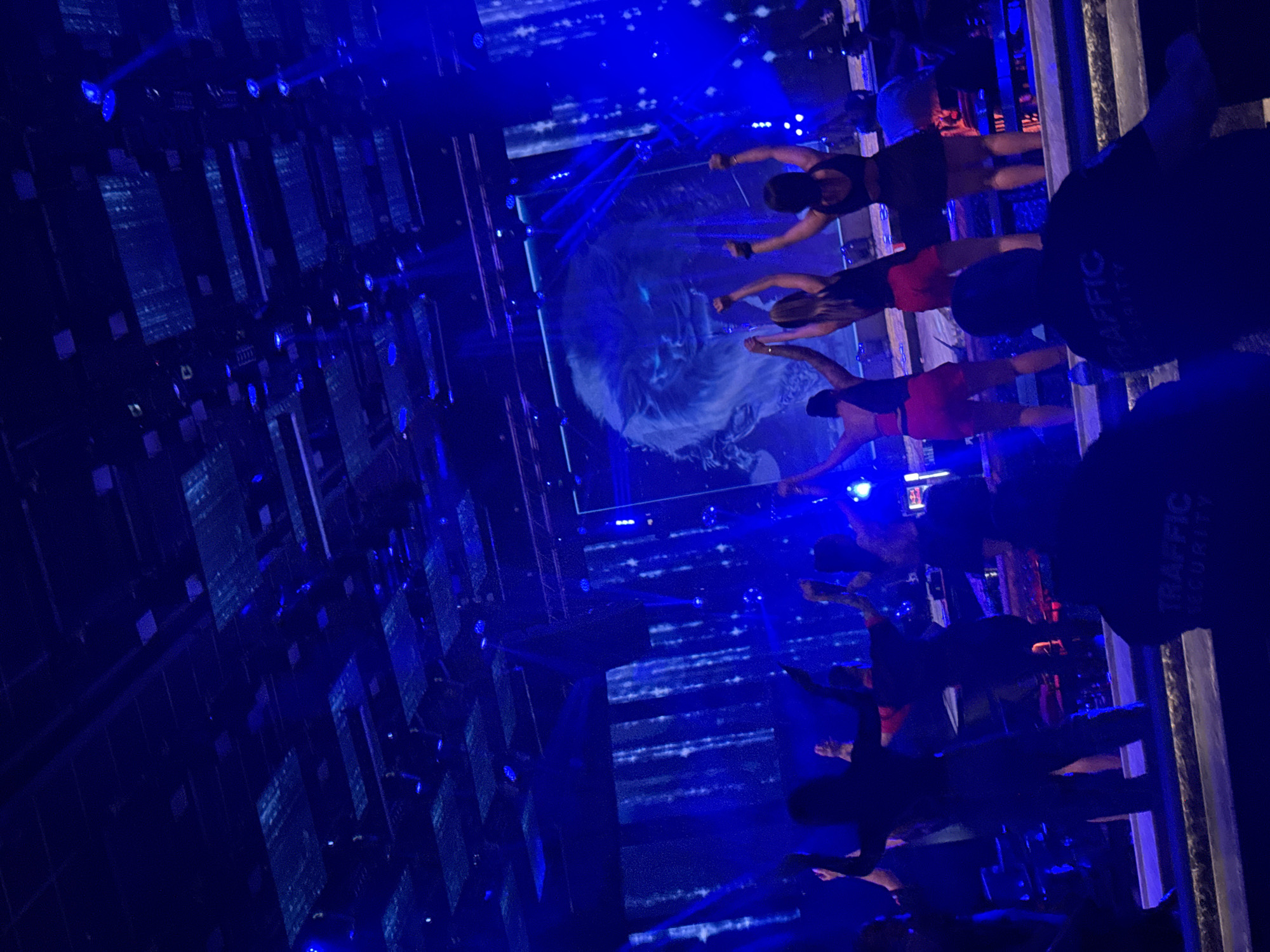 The image captures an indoor scene within the Traffic Club, Plovdiv, Bulgaria, characterized by predominant blue artificial lighting. In the foreground, two individuals, seen from behind, wear dark shirts with "TRAFFIC SECURITY" printed in light text on their backs, indicating their role as security personnel. They are positioned below a raised stage or platform.
On this stage, approximately 6-8 individuals are visible, mostly women, engaging in a performance or dancing. Four women are prominently featured in the midground, seen from behind with their arms raised in the air. Their attire includes dark tops, with two wearing red shorts and one wearing a dark skirt or shorts. Other figures are partially visible further back on the stage, including one seated near what appears to be a DJ booth or sound equipment on the far right.
The stage's background is dominated by large LED screens. A central screen displays a glowing, stylized lion's head with bright blue eyes, while vertical screens on either side show dynamic, abstract blue light patterns resembling falling particles. The ceiling above features a dark, grid-like structure with embedded blue lights. The scene suggests a lively nightclub or concert event.
