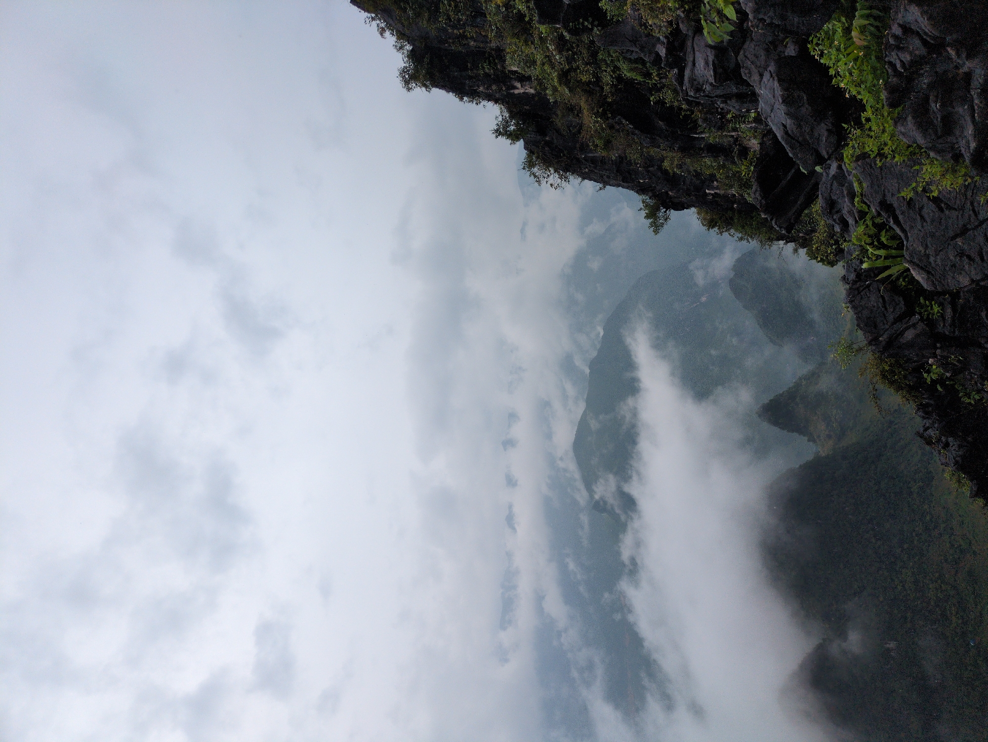 The image captures a dramatic mountain landscape in Đồng Văn Commune, Vietnam, under a cloudy sky. In the foreground, a rugged, dark rocky cliff face with sparse green vegetation juts out from the right side of the frame. Beyond this, a vast expanse of mist-shrouded mountains stretches into the distance. The valleys between the peaks are filled with thick white clouds, creating a sense of depth and mystery. The overall impression is one of a remote and wild natural environment. There are no people or man-made objects visible in the image.