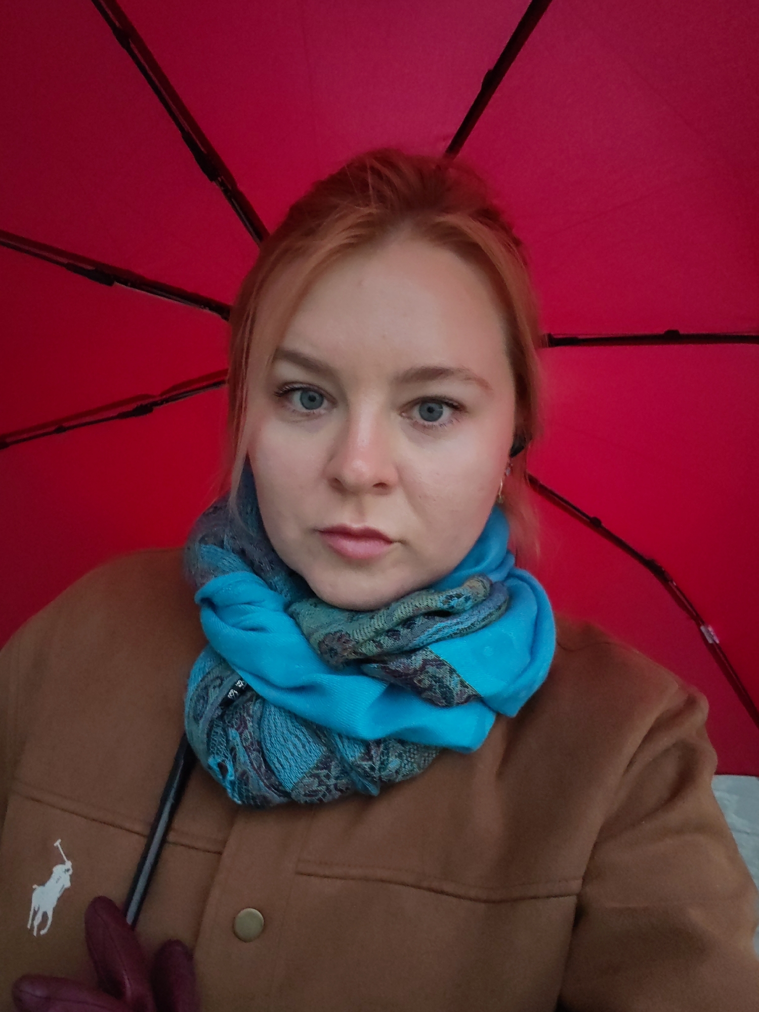 A woman with light blonde hair and blue eyes is centered in the frame, wearing a brown jacket, a blue and teal patterned scarf, and what appears to be a red umbrella. A white polo logo is visible on the jacket. The background is a solid red color, suggesting the interior of the umbrella. It is not possible to determine the location in Izhevsk, Russia, based solely on this image.
