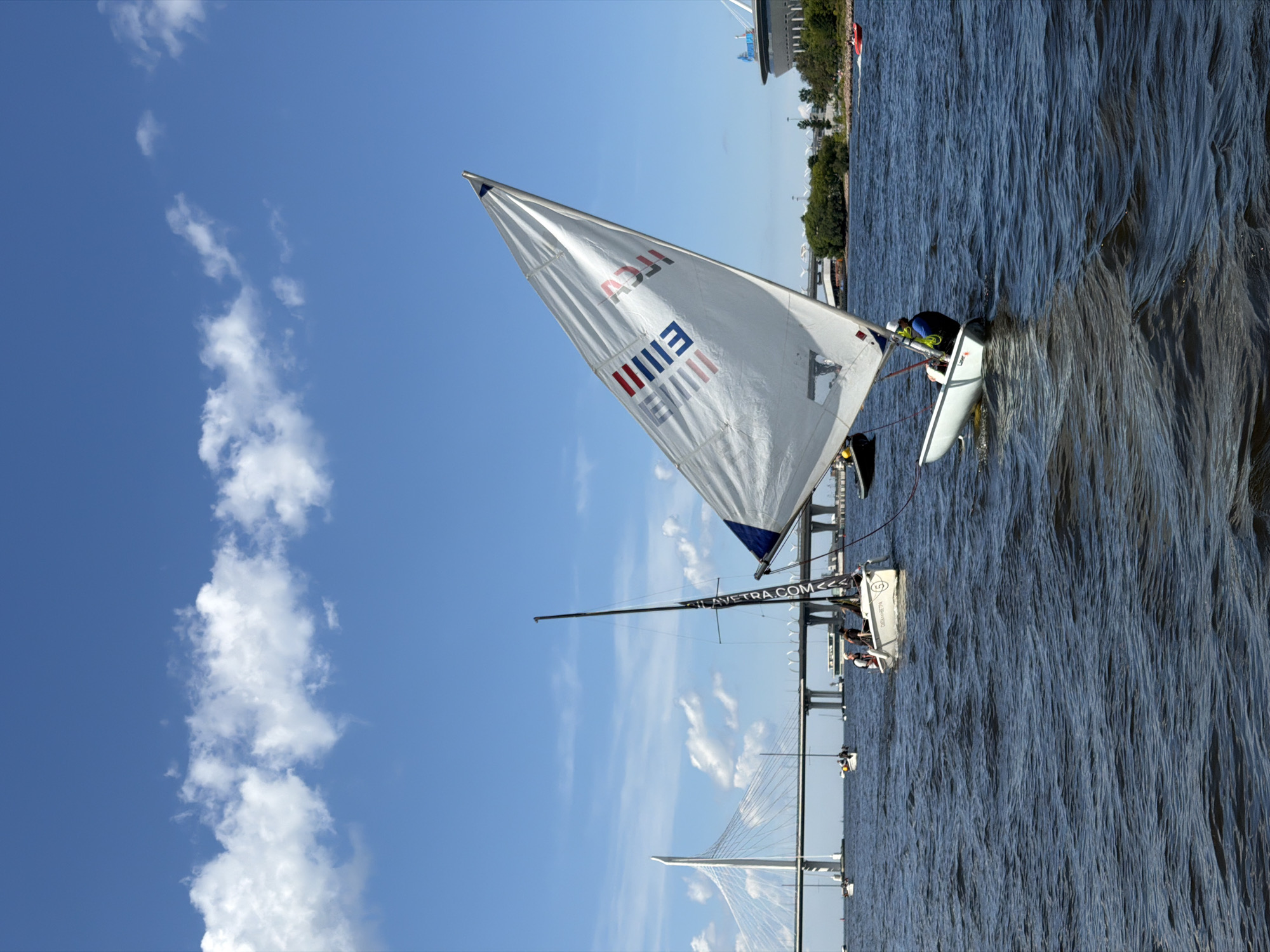 A single person is sailing a small white sailboat on a body of water. The sailboat has a white sail with red and blue stripes and lettering. The person is wearing a blue shirt and a white hat. In the background, there is a large suspension bridge and a stadium. The sky is blue with some white clouds. The water is dark blue and choppy. The image appears to be taken on a sunny day. The location is likely St. Petersburg, Russia, given the presence of the stadium and bridge which resembles the Gazprom Arena and the Western High-Speed Diameter.