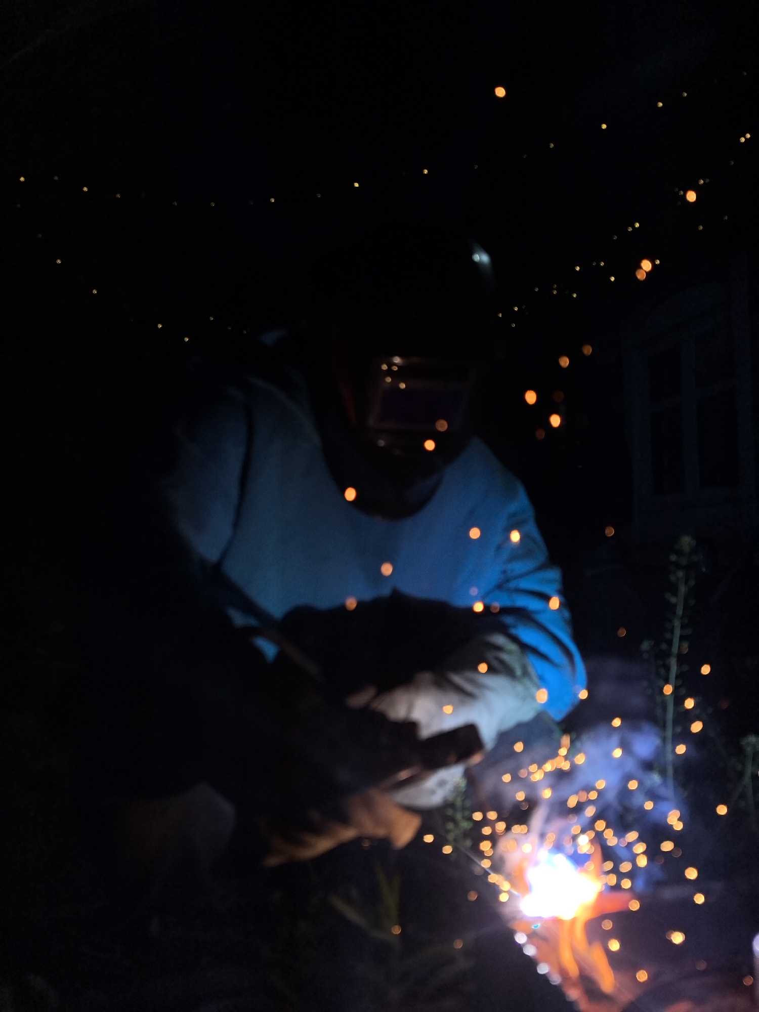 A person wearing a welding helmet, blue sweatshirt, and gloves is welding in the dark. Sparks fly from the weld. The background is dark, with some blurred light points and a window-like structure visible in the distance. The location is likely outdoors, with some plant life visible. The city of Noviy, Russia is not directly evident from the image.