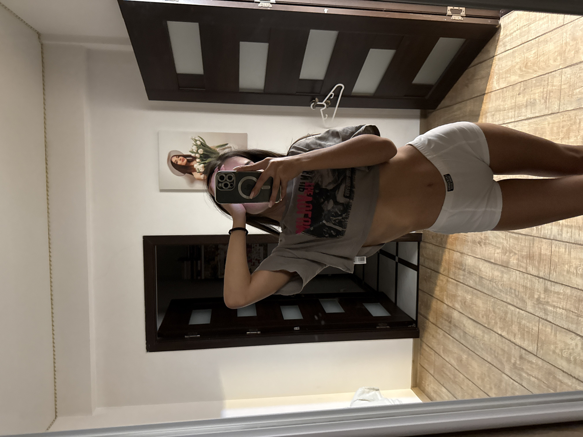 A young woman is taking a selfie in a hallway, reflected in a large mirror. She is wearing a grey cropped t-shirt and white boxer briefs. Her right arm is raised behind her head, and she is holding a smartphone with her left hand to capture the image. In the background, through a doorway to her left, a bedroom is partially visible, with a tiled floor and a bed. To her right, another doorway leads into a different room, also with a tiled floor. A framed picture of a woman wearing a hat and holding a bouquet of flowers hangs on the wall above the first doorway. The hallway has wooden flooring and white walls. The scene appears to be indoors, likely in a residential setting, potentially in Chișinău, Moldova, as suggested by the information provided.
