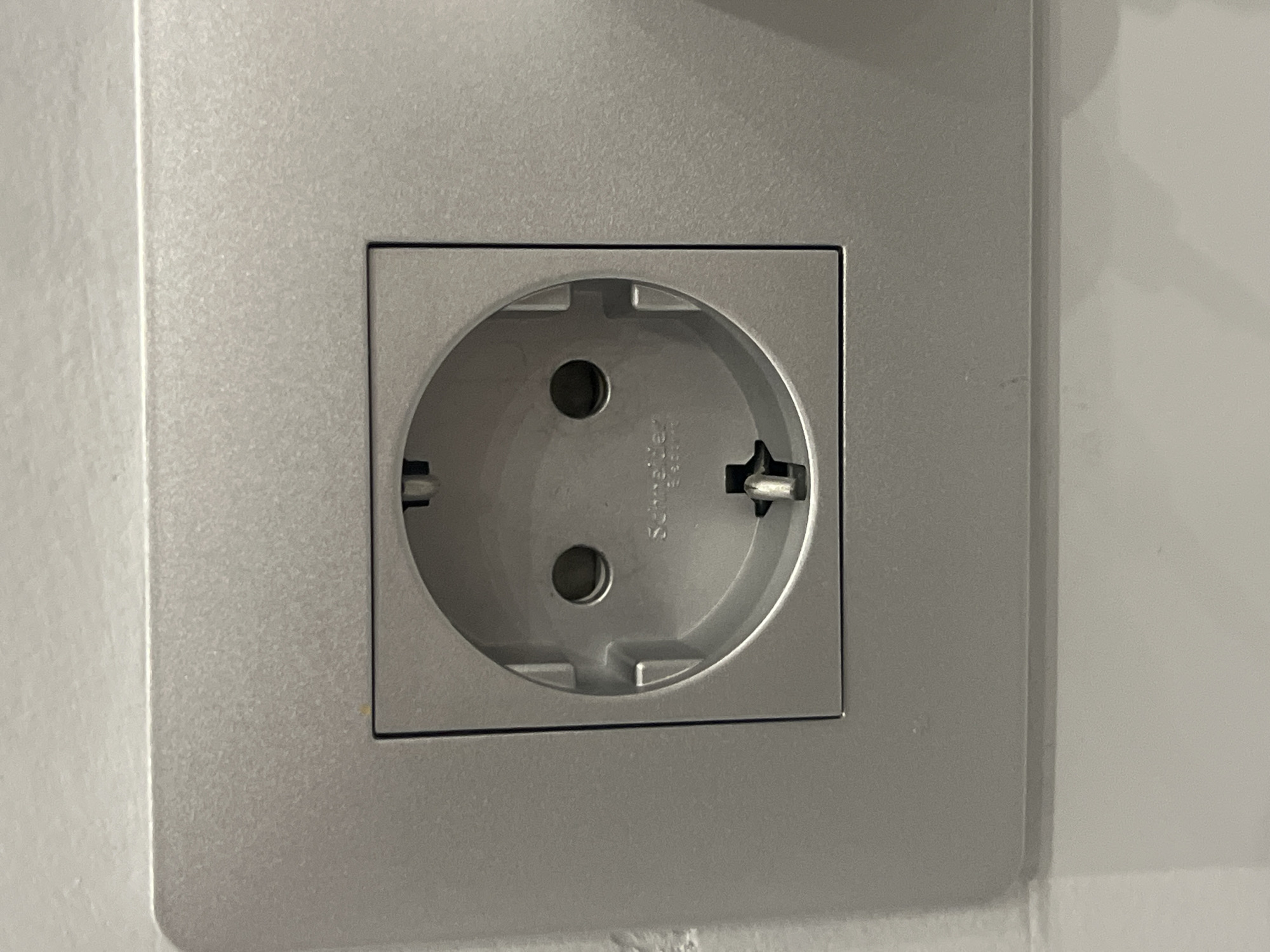 A close-up, eye-level, medium shot shows a light gray, square electrical outlet installed on a white wall. The outlet is a standard European type with two round holes and a grounding slot. The outlet plate is flush with the wall and has a subtle brushed metal texture. The background wall is painted white and appears to be slightly textured. There are no people or other objects visible in the frame. The image is well-lit and in focus. This image was taken in Podgumer, Bulgaria.