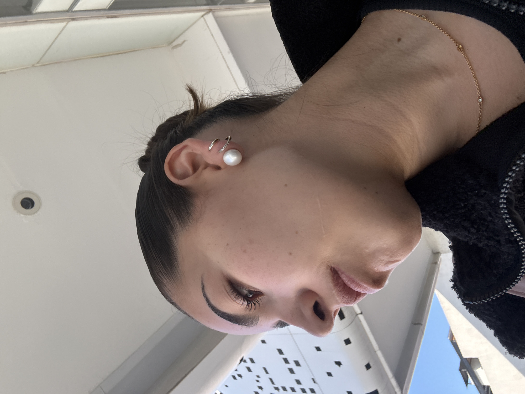 The image is a close-up portrait of a woman, depicted in profile and oriented on her side, looking downwards and slightly to the right. Her dark hair is styled back, emphasizing her meticulously groomed eyebrows and prominent, long eyelashes. She is adorned with striking jewelry: a large, radiant pearl earring paired with a delicate, possibly diamond-studded, double cuff on her left ear. Around her neck, a fine gold chain necklace with small beads and a tiny diamond pendant is visible. She wears a dark, textured garment, likely a black fleece or boucle jacket, which frames her neck.
The background reveals a bright outdoor setting, indicated by a clear blue sky on the right. Modern architectural elements surround her. To her right, a light-colored structure, possibly an overhang or ceiling, contains a small black circular light fixture. Below this, a contemporary white building facade with numerous small, dark square cutouts suggests an urban environment. Another angular, light-colored building is partially visible on her left. The scene captures the woman posing for a photograph, highlighting her features and accessories in daylight. The setting is consistent with a modern urban area, located in Puebla, Mexico. No specific activity other than posing is taking place, and no text is visible.