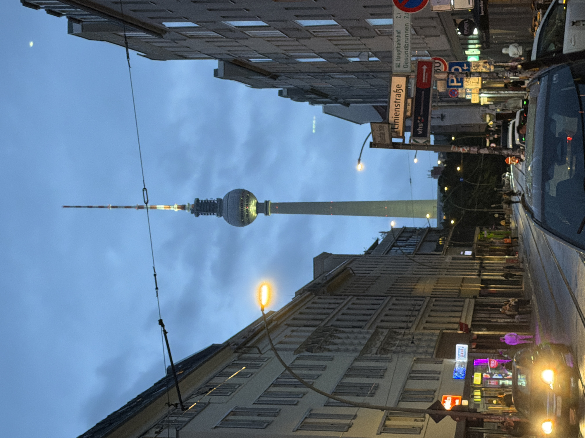 The image depicts a street scene in Berlin, Germany, at dusk. The prominent Fernsehturm (Television Tower) stands tall in the background, illuminated against the twilight sky. The street is lined with buildings, some with visible windows and architectural details. Streetlights cast a warm glow on the pavement. Cars are visible on the street, one in the foreground with its headlights on, and others further down the road. Pedestrians are present on the sidewalks, some walking and others gathered. Several street signs are visible, including one indicating "Linienstraße" and another pointing towards "Hauptbahnhof" and "Gesundbrunnen". The overall atmosphere suggests a typical urban evening in the city.