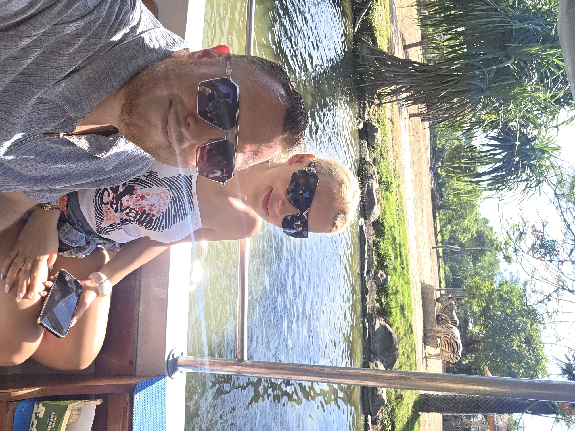 A man and woman are on a boat next to an enclosure of zebras. The woman holds a mobile phone. Đà Nẵng, Vietnam cannot be determined from the image.