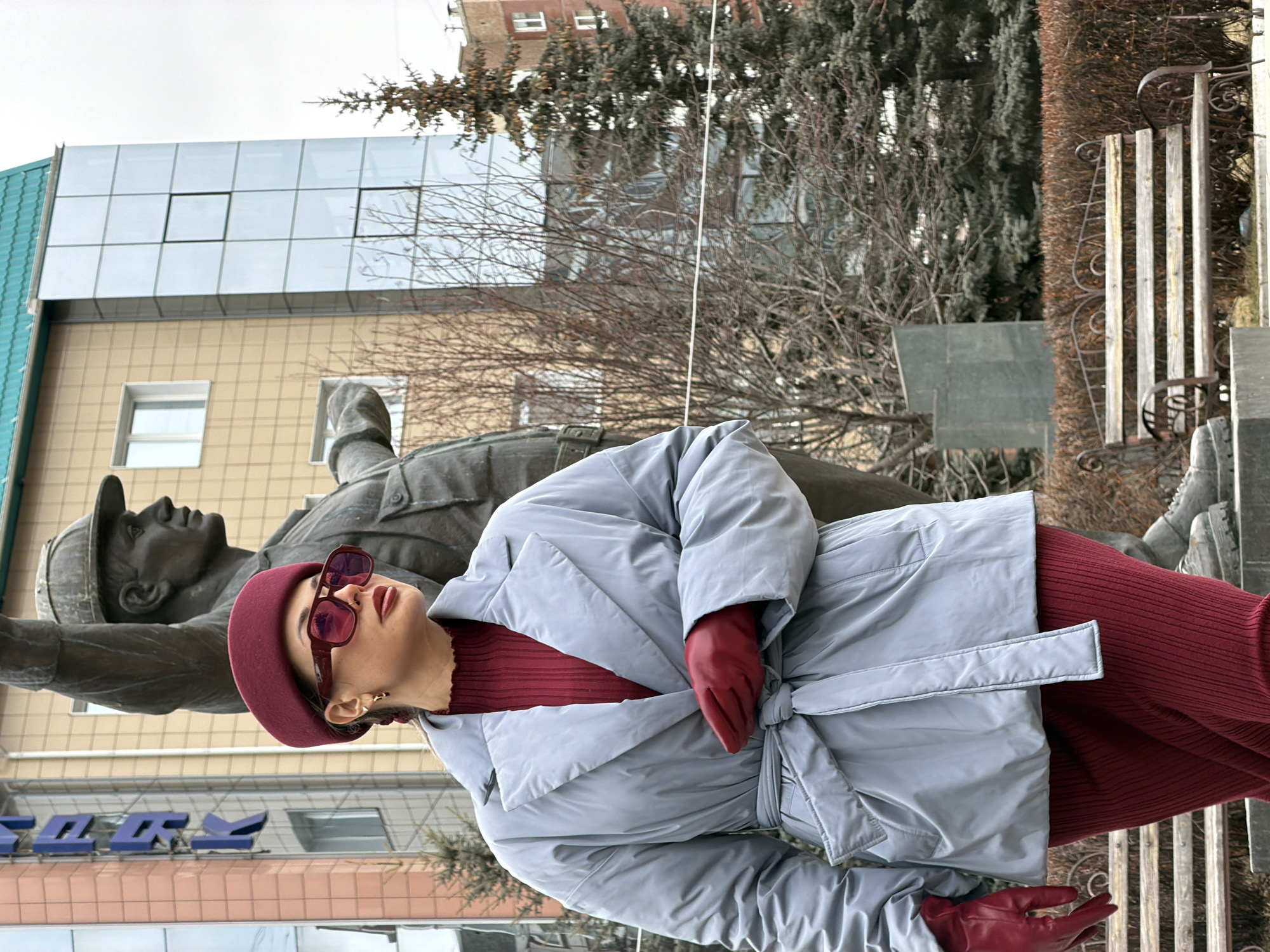 The photograph depicts a woman posing in front of a monument in what appears to be an urban setting. The woman is wearing a burgundy hat, sunglasses, a burgundy turtleneck sweater, a light blue, belted coat, and burgundy gloves. She stands to the right of a weathered bronze or stone monument depicting two figures, likely laborers or soldiers.
In the background, there's a modern building with a glass facade, as well as older brick buildings and bare trees, indicating it might be late autumn or winter. To the right of the monument, there is a park bench.
Based on the context clues, the location is Krasnoyarsk, Russia. The monument is likely a local landmark or memorial. The time of day seems to be during daylight hours, judging by the natural lighting.