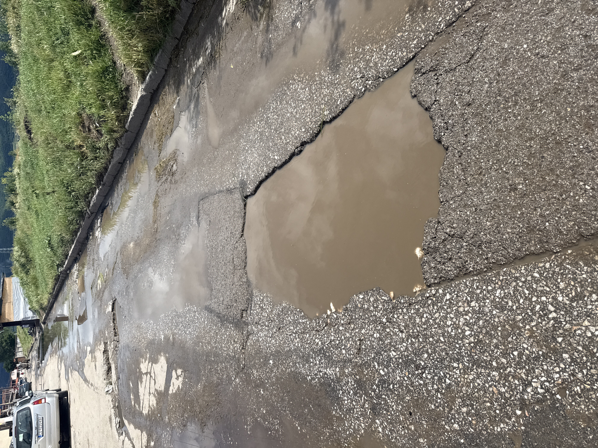 The image displays a severely damaged asphalt road characterized by numerous large potholes filled with murky, brown water. The road surface is heavily degraded, with chunks of asphalt missing and cracked areas, revealing the underlying aggregate.
On the left side of the road, a lush green, overgrown grassy verge slopes upwards, suggesting a natural embankment or hillside. In the distance beyond the verge, mountains or large hills are faintly visible. A concrete curb or divider runs along the edge where the grass meets the road.
The muddy water in the potholes reflects the bright sky and clouds, indicating it is daytime, possibly with sunny or partly cloudy weather. Shadows from unseen objects, likely trees or utility poles, are also visible reflecting in the water on the right side of the road.
Further down the road on the left, a silver car is parked, partially visible. Its license plate clearly shows the text "GO 9224 KB". Beyond the car, some construction-related materials, possibly wooden planks and a white tarpaulin, can be seen, hinting at ongoing work or an informal setup. The overall scene depicts a neglected road in a semi-rural or semi-urban environment, with standing water from recent rainfall or poor drainage.