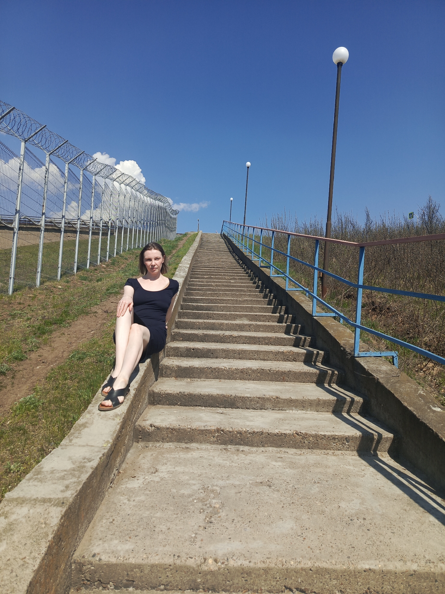 A woman is sitting on the concrete edge of a long staircase, under a bright blue sky with a few clouds. The staircase stretches upwards, bordered on one side by a fence topped with barbed wire and on the other side by a blue railing with lamp posts. Brown vegetation is visible beyond the railing. The woman is wearing a dark dress and sandals, and is looking directly at the camera. This scene is located on the stairs leading to the "Monument to the First Tent," in Chaikovsky, Russia.